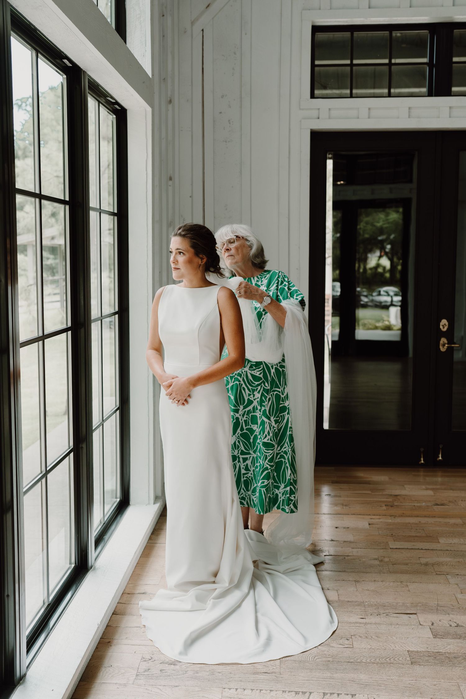 Wedding scene with a bride in white gown and guest in green floral dress by sunlit windows.