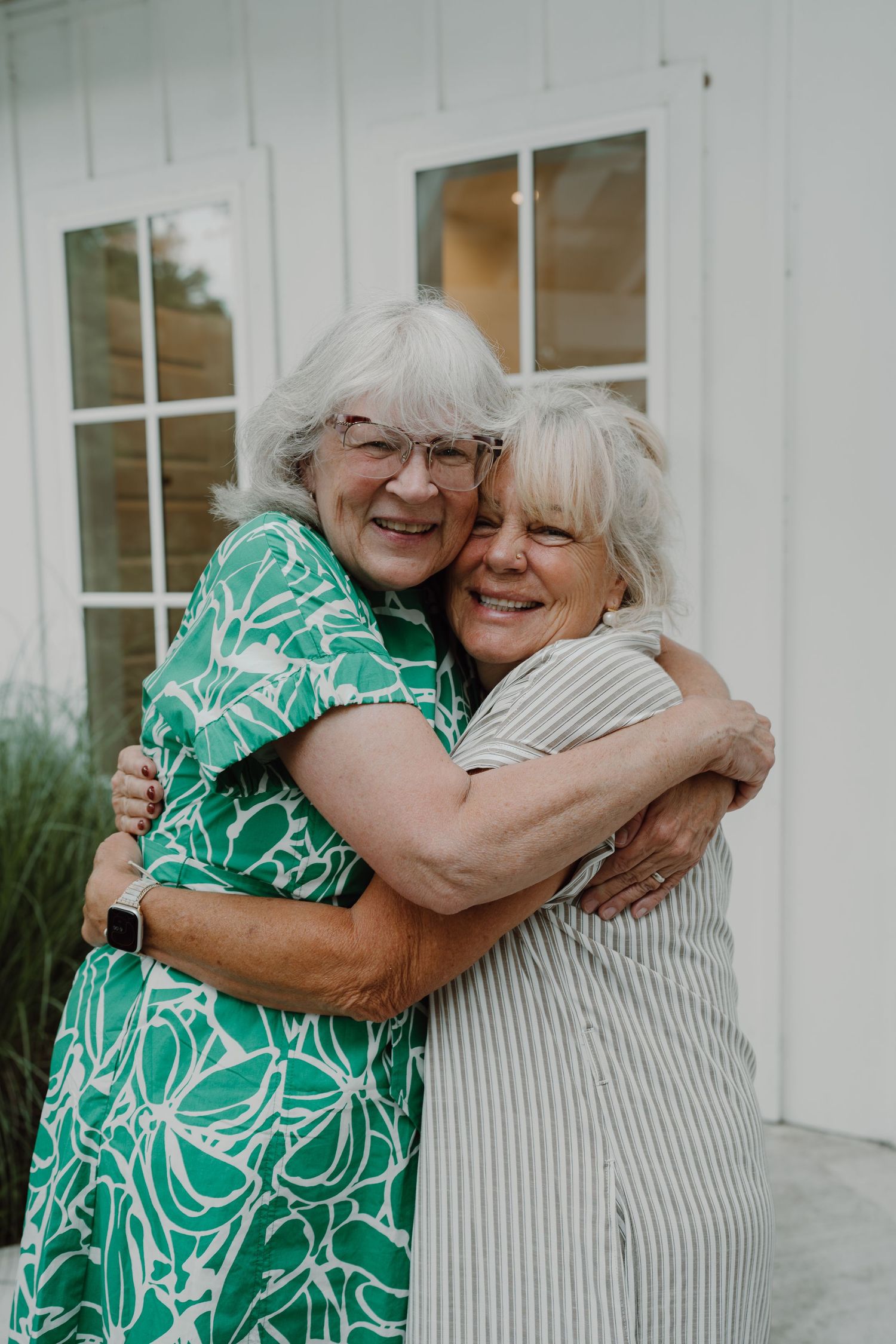 Two women embracing and smiling outside a white building during a special occasion.