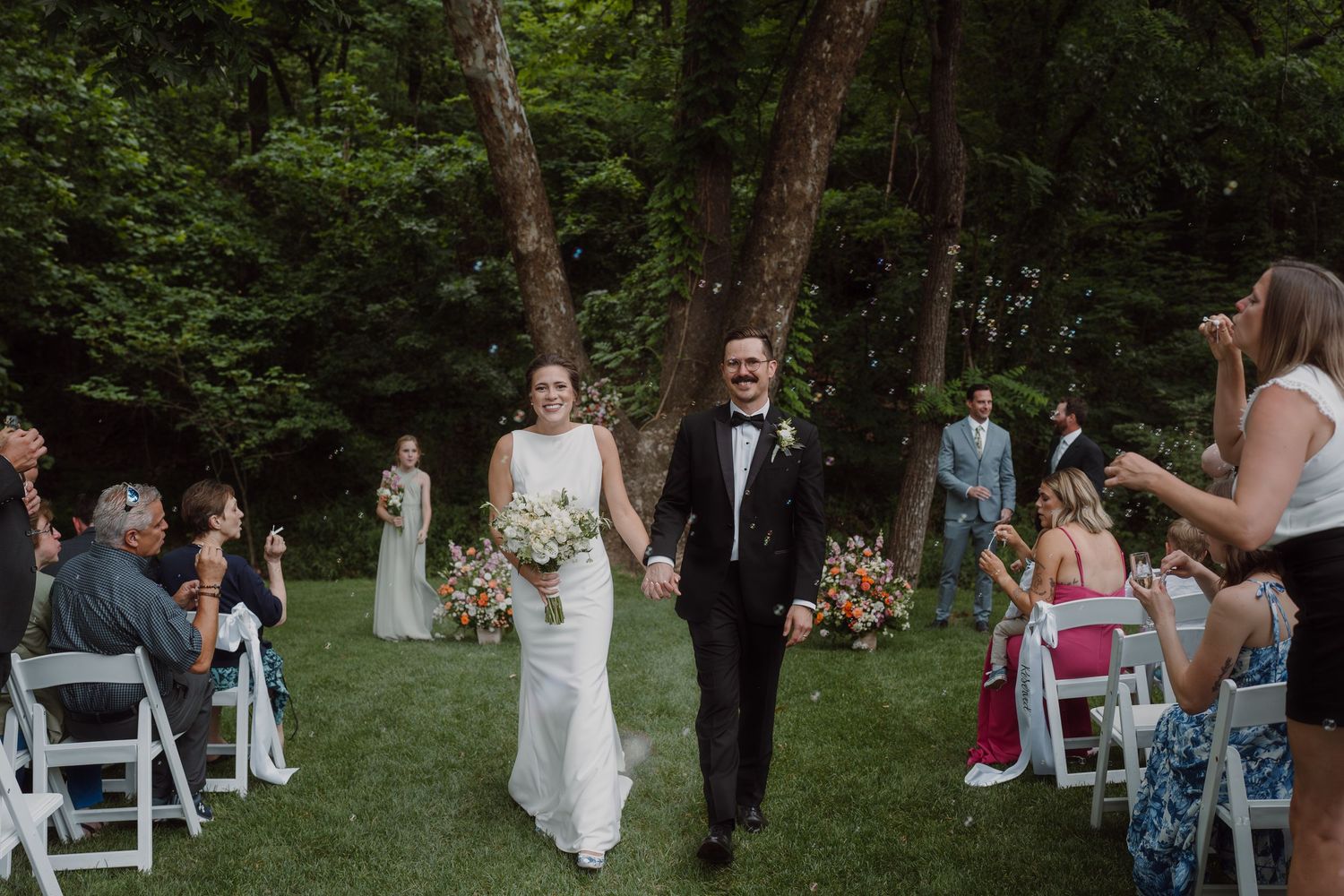 Outdoor wedding ceremony with white chairs and guests gathered in a wooded setting.