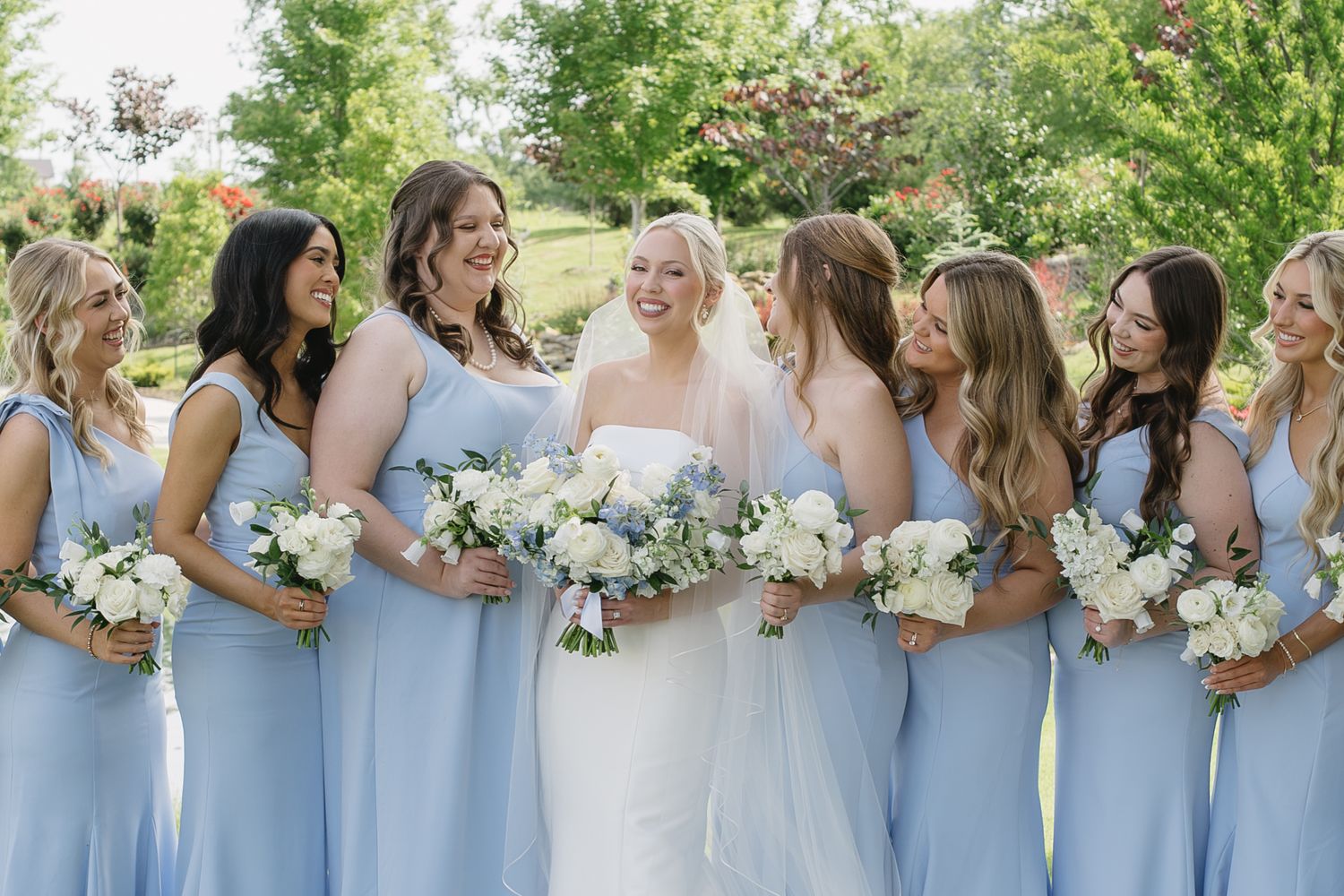 Bride in white gown surrounded by bridesmaids in light blue dresses holding white floral bouquets outdoors.