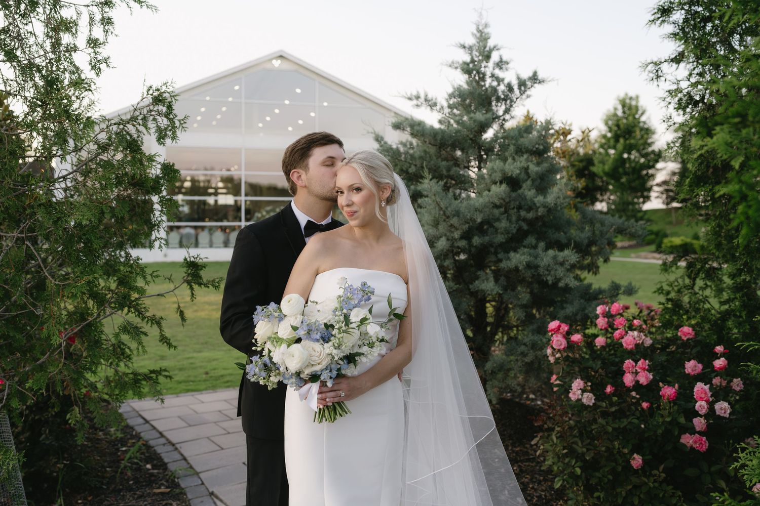 Bride and groom pose in garden venue with white bouquet of hydrangeas, roses, and greenery at outdoor wedding reception.