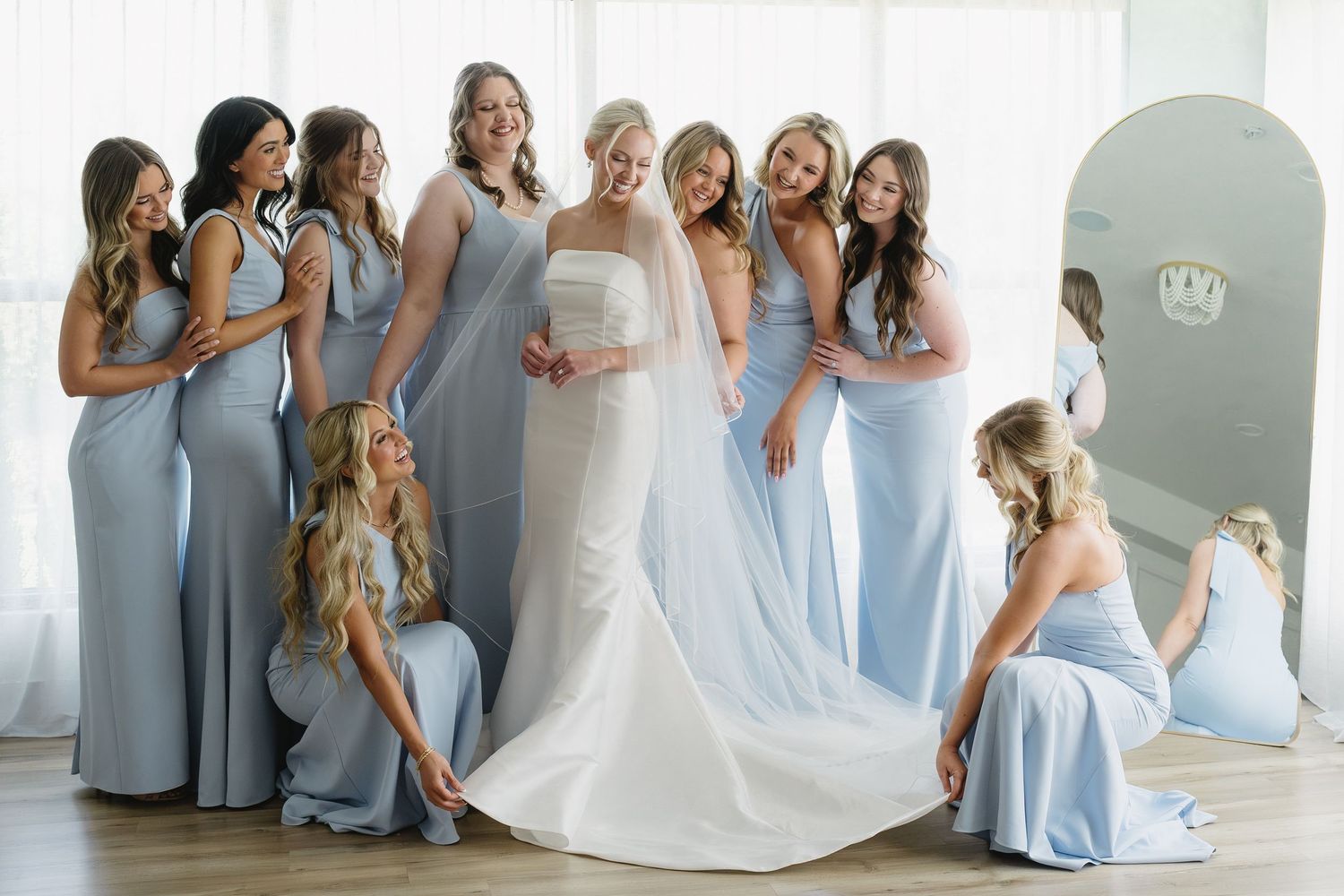 Bride in white gown surrounded by smiling bridesmaids in light blue dresses, posing together in a bright elegant room.