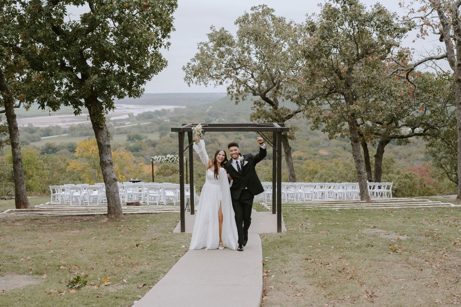 Newlyweds celebrate under a rustic wooden arbor at an outdoor hilltop ceremony venue with white chairs.
