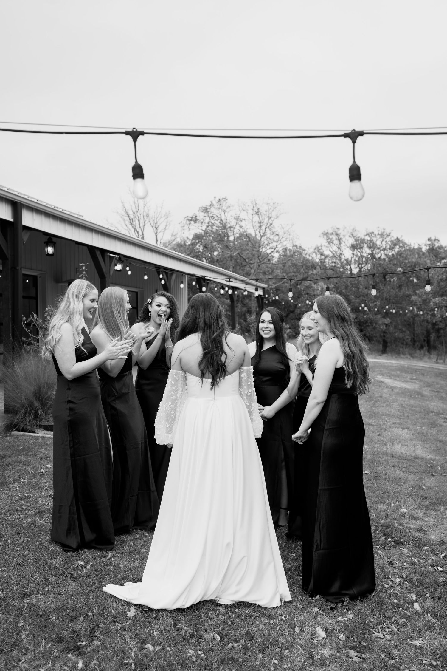 Black and white photo of wedding party gathered outside venue with string lights overhead.