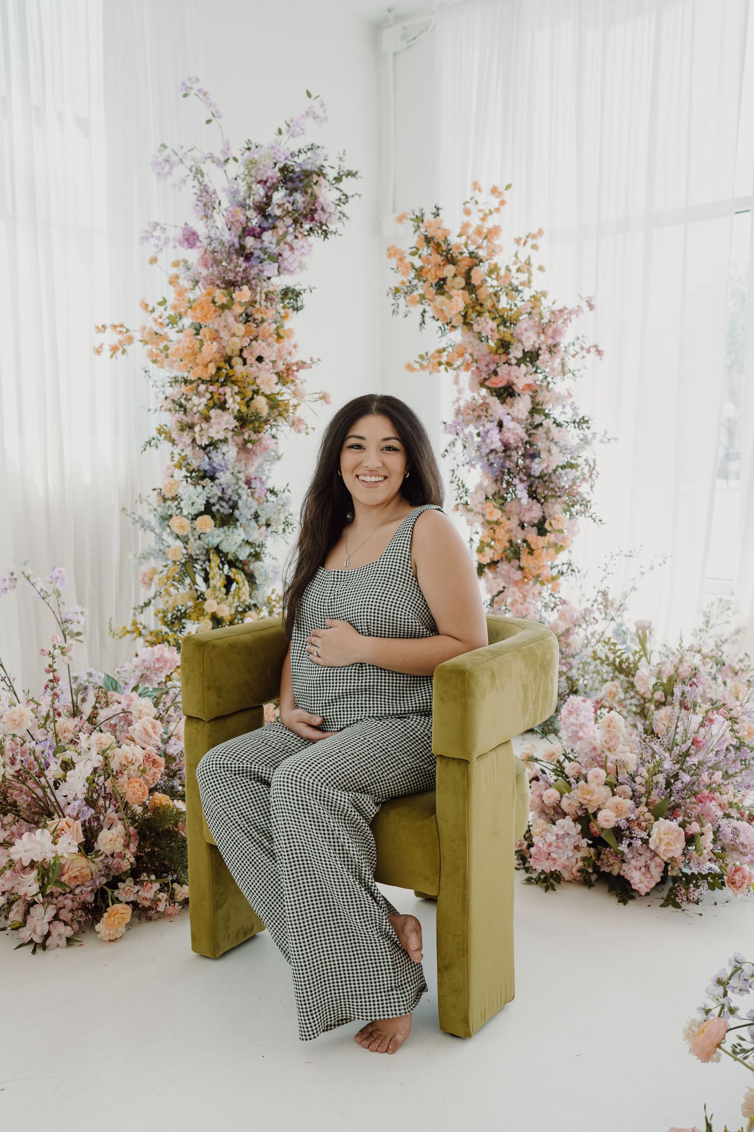 A maternity photoshoot in a bright white studio with floral arrangements and a green velvet chair.