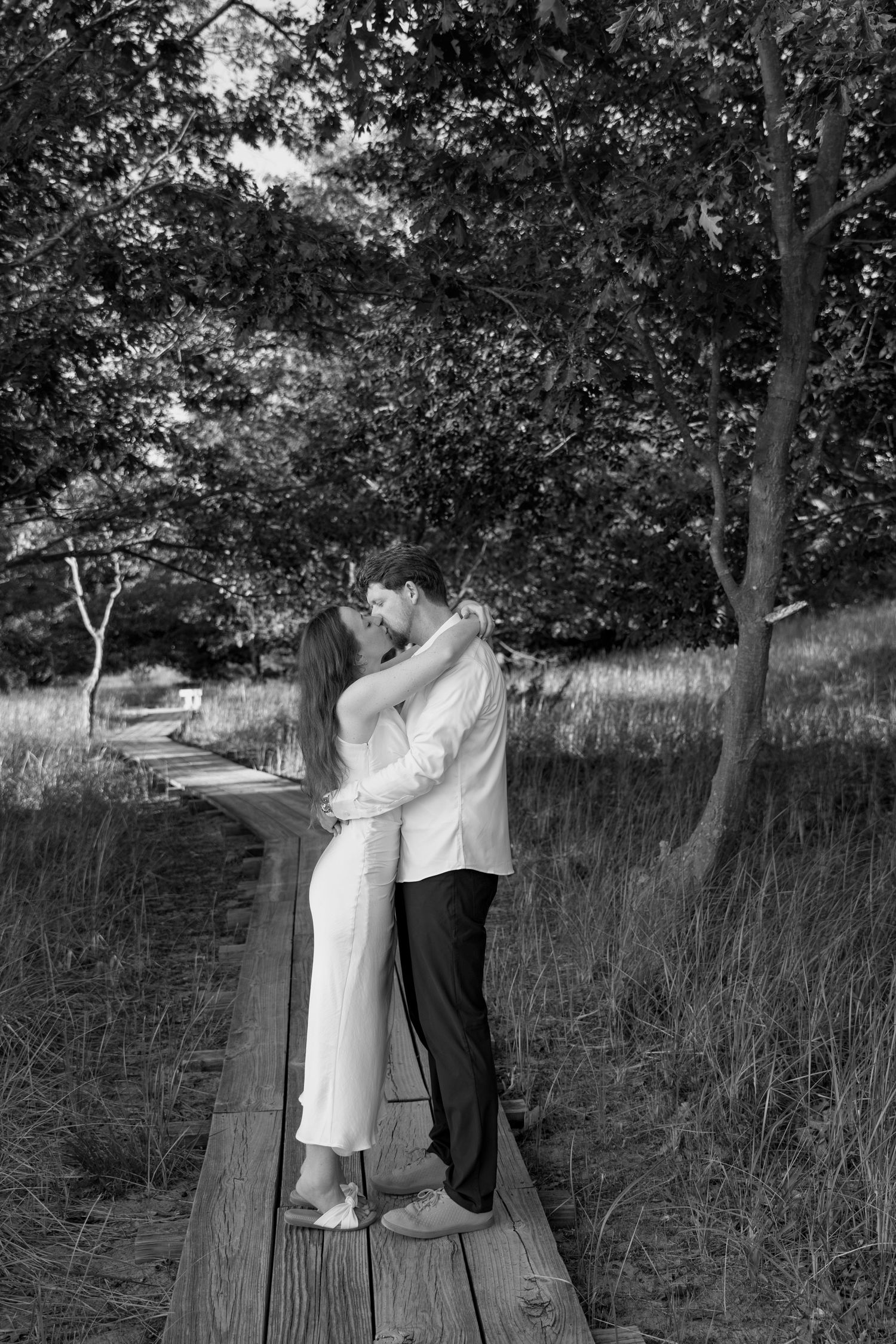Black and white photo of a couple embracing on a wooden boardwalk surrounded by tall grass.