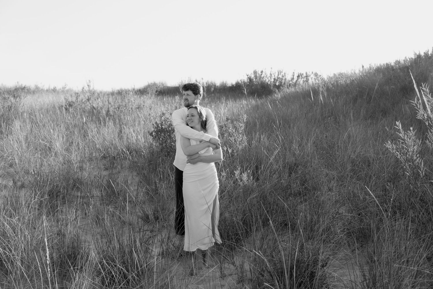 A couple embraces tenderly on a grassy sand dune during golden hour.