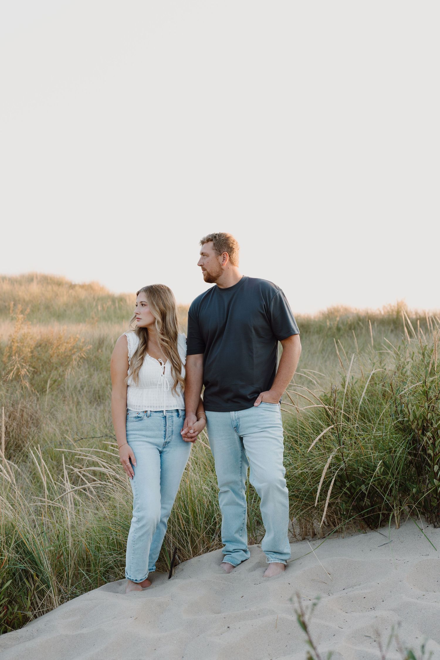 Two people stand together on a sandy hill surrounded by tall beach grass at dusk.