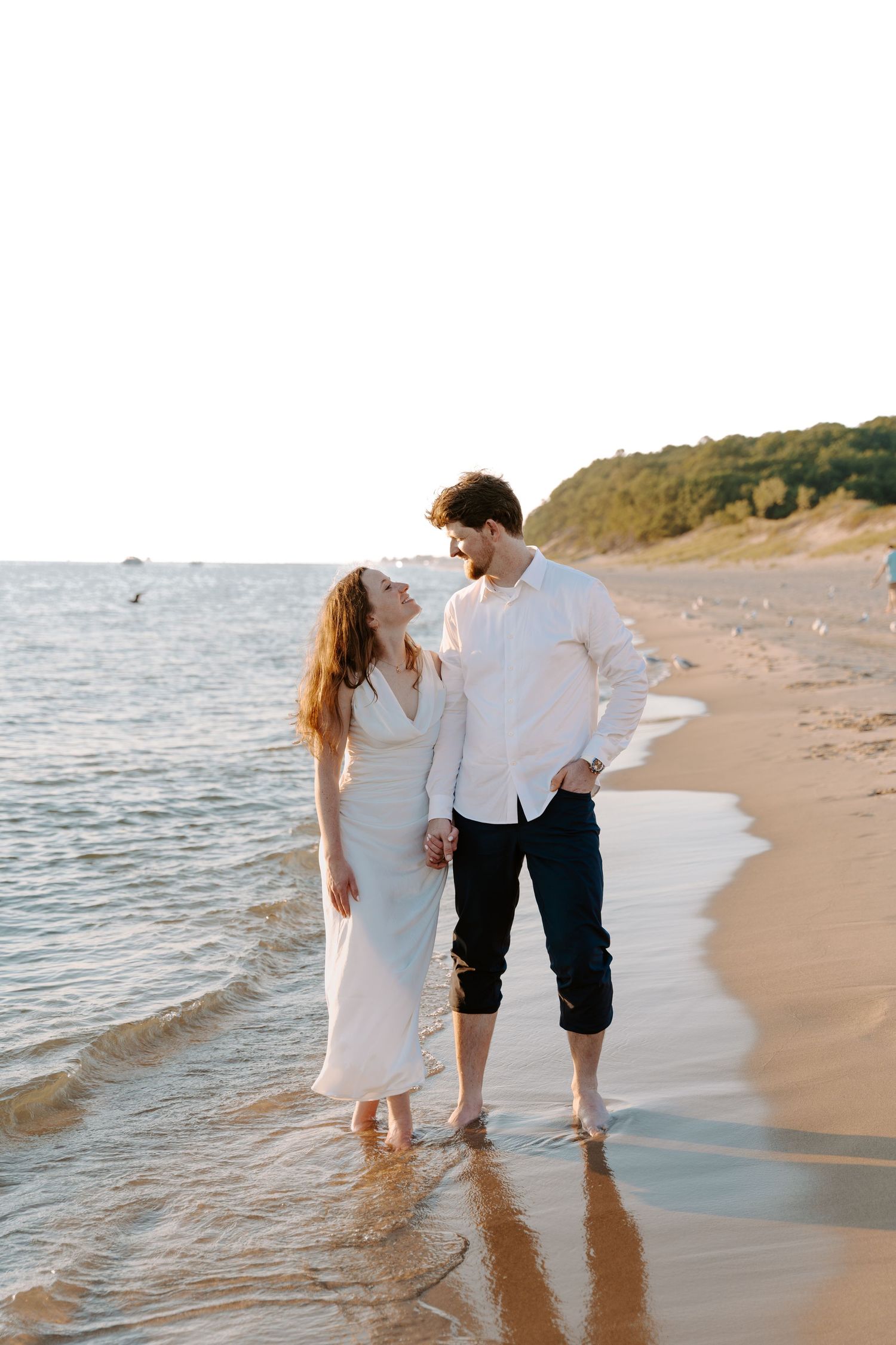 Couple walking barefoot in shallow ocean water along scenic beach.