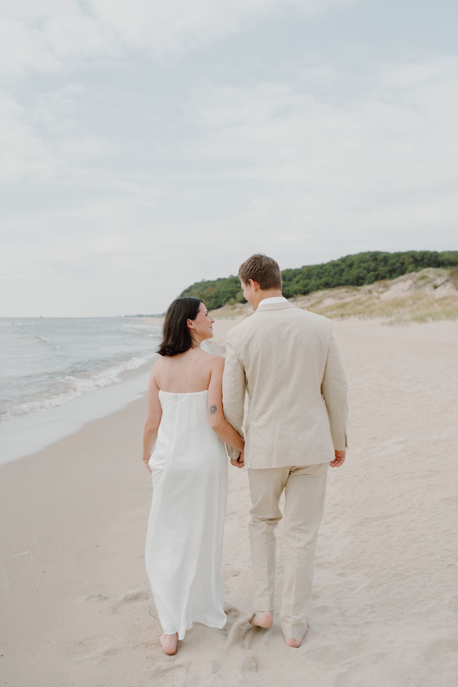 A couple strolls barefoot along a pristine beach with sand dunes and ocean views.