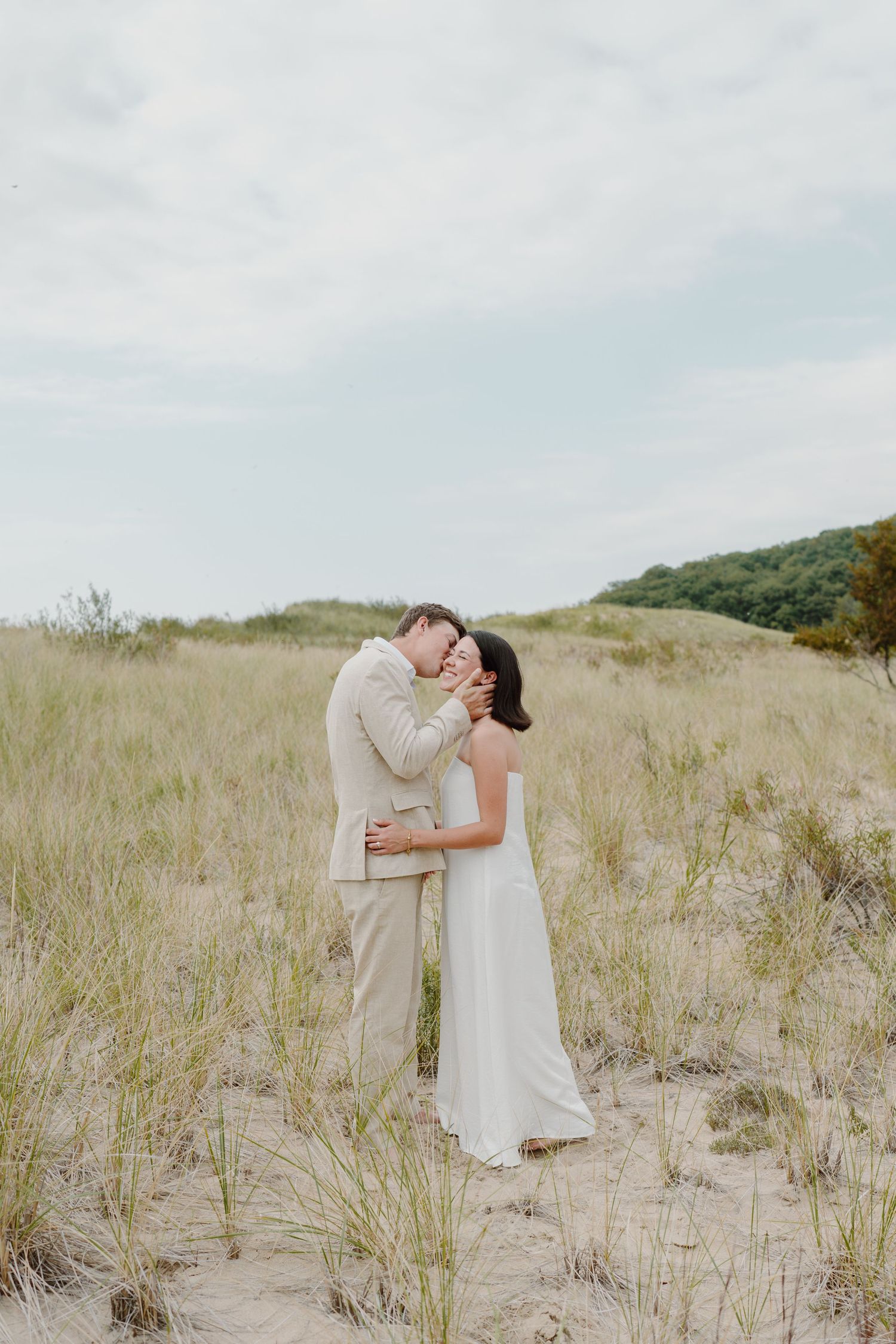 Candid moment of newlyweds sharing a tender embrace among coastal dunes on a cloudy day.