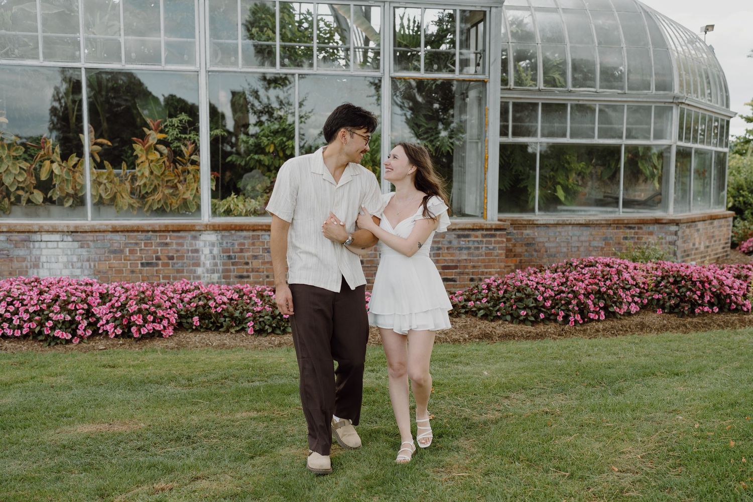 A couple poses playfully outside a glass conservatory surrounded by lush gardens.