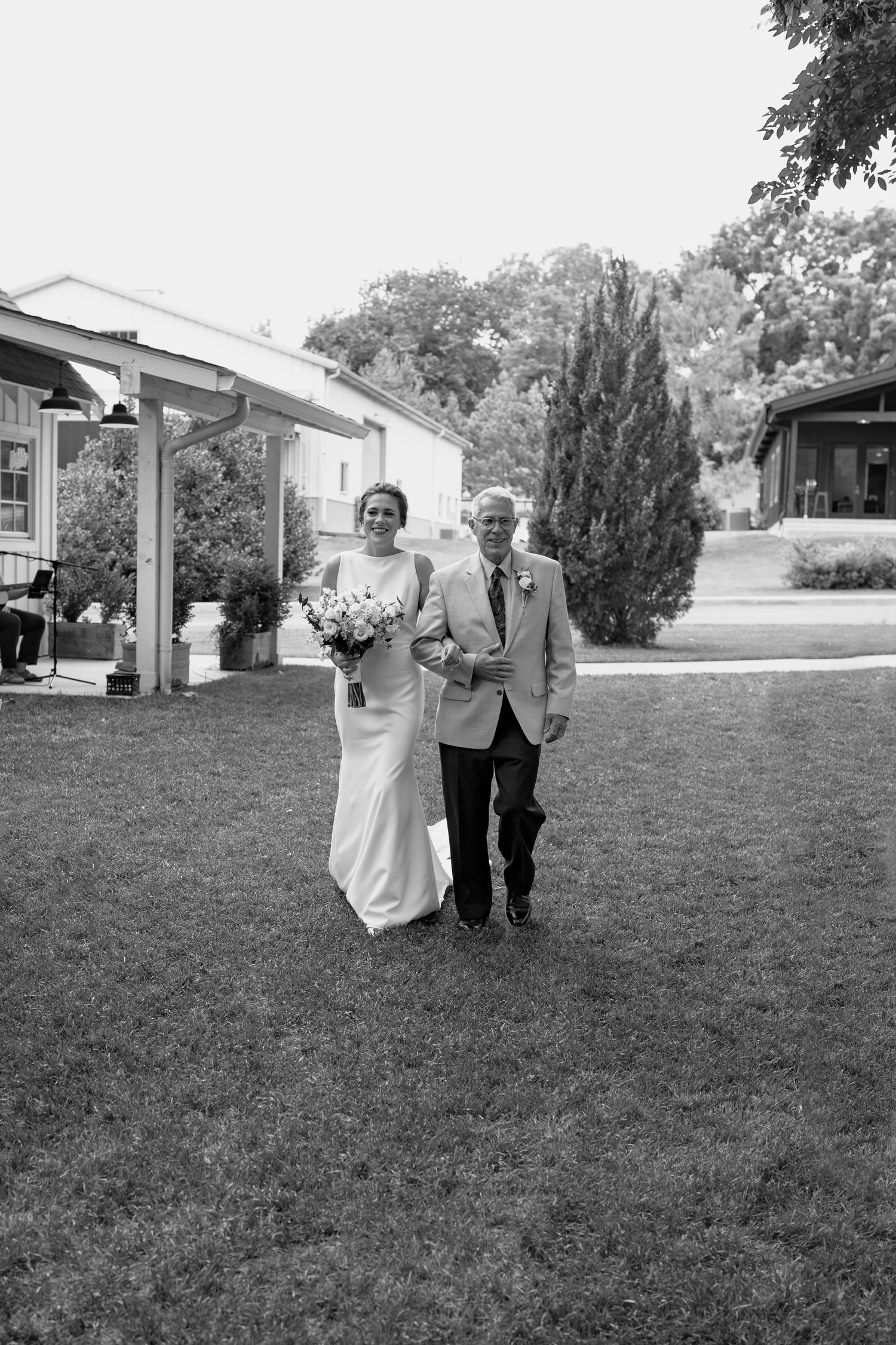 A black and white wedding photo shows a couple walking across a lawn with buildings and cypress trees.