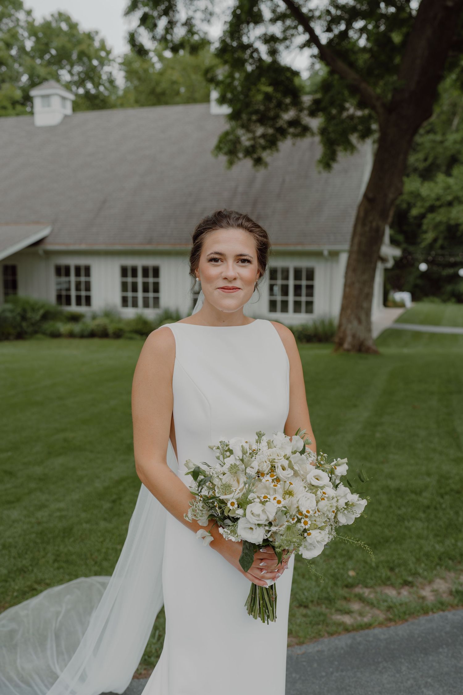 A bride poses with her white floral bouquet against a backdrop of green lawn and white building.
