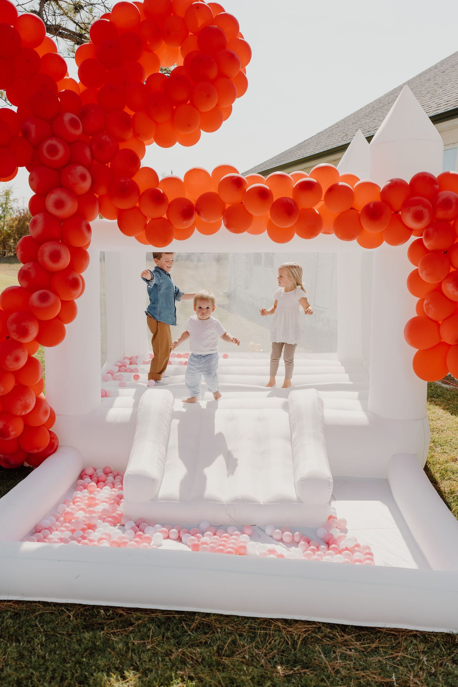 Children enjoying a white bounce house with red balloon decorations at an outdoor party.