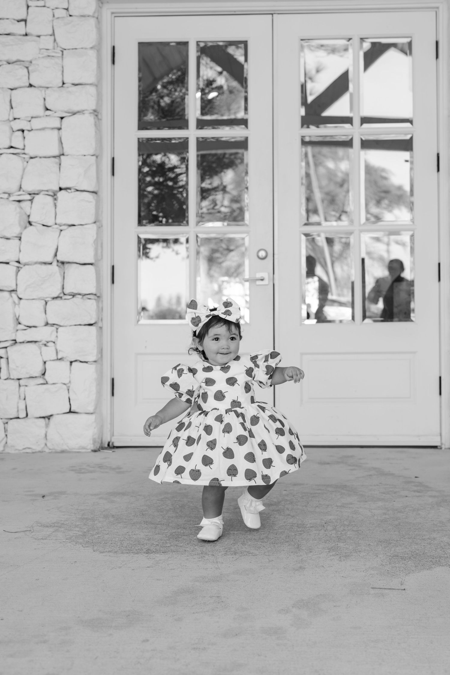 Black and white image of toddler in patterned dress playing near building entrance.