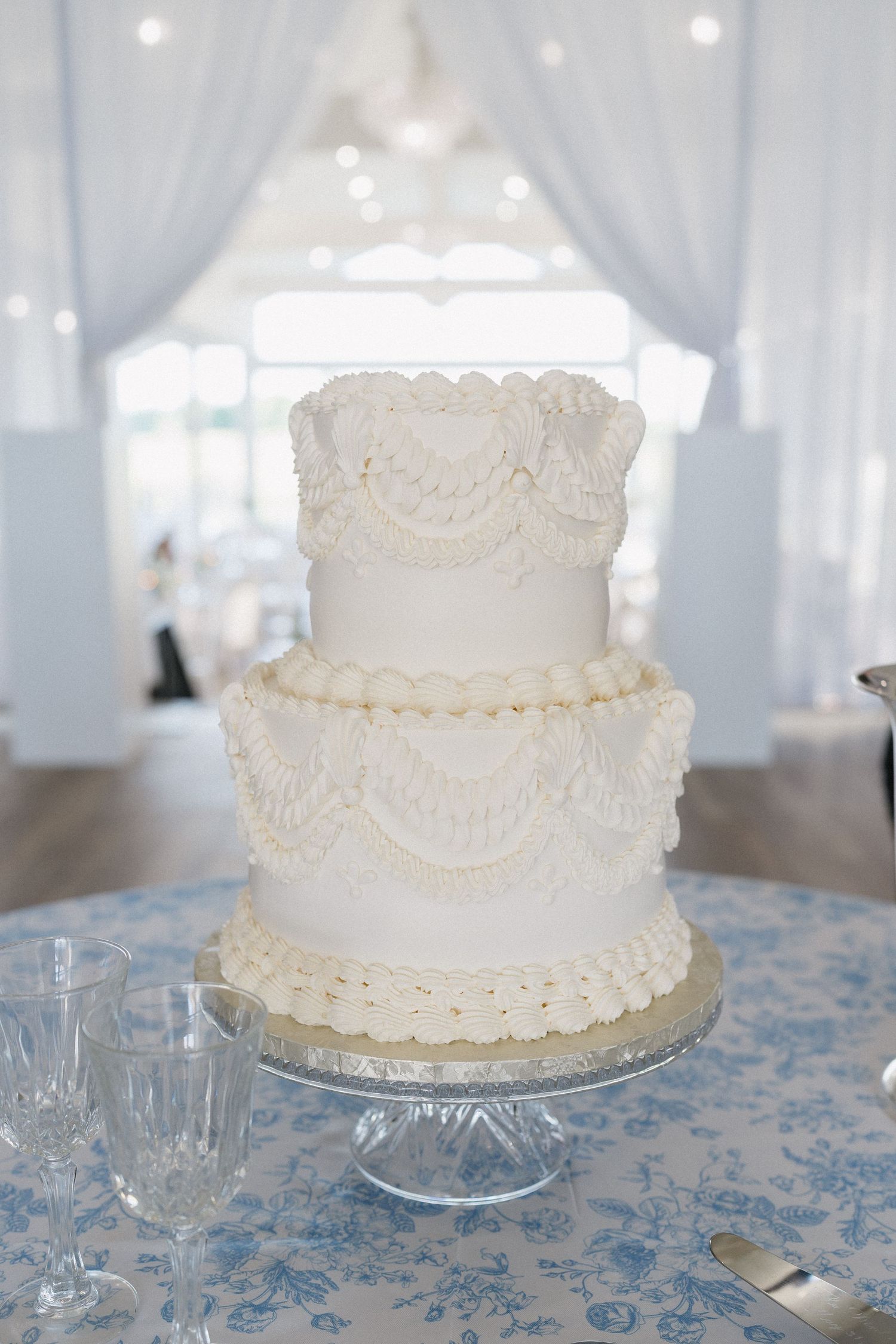 Elegant three-tier white wedding cake with intricate lace piping on a crystal stand in a bright white reception venue.