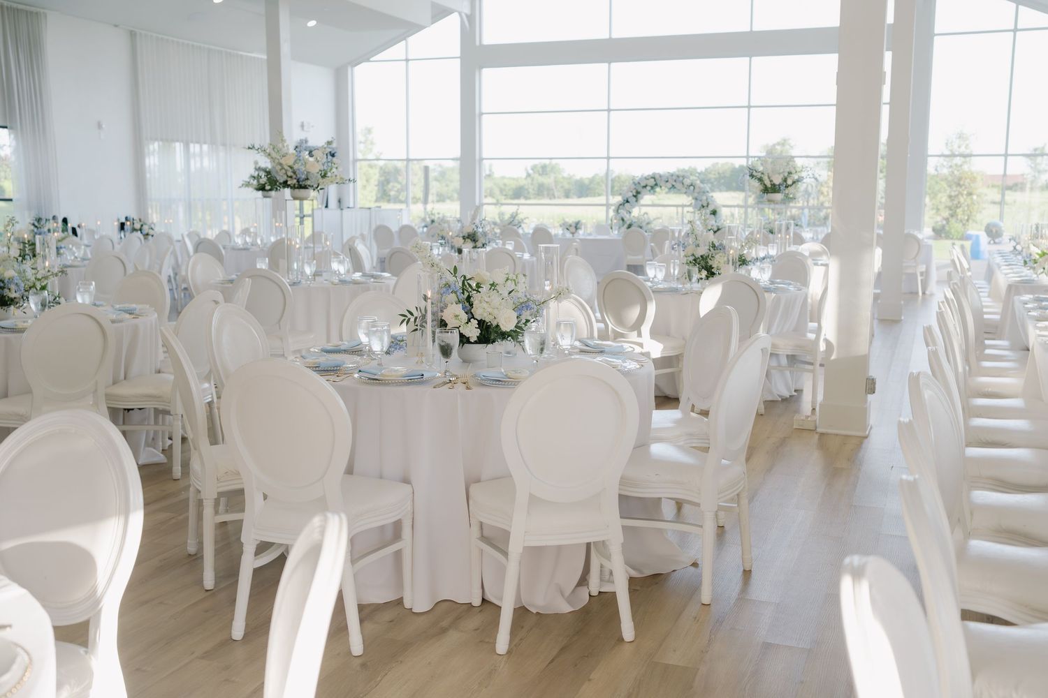 Elegant white wedding reception hall with round tables, floral centerpieces, and large windows offering a bright, airy atmosphere.