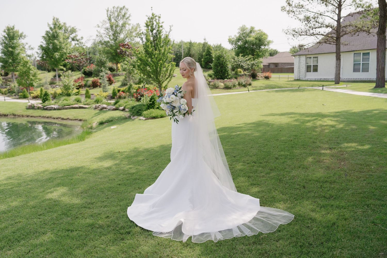 Bride in elegant white gown with long veil holding bouquet on lush green lawn near scenic pond.