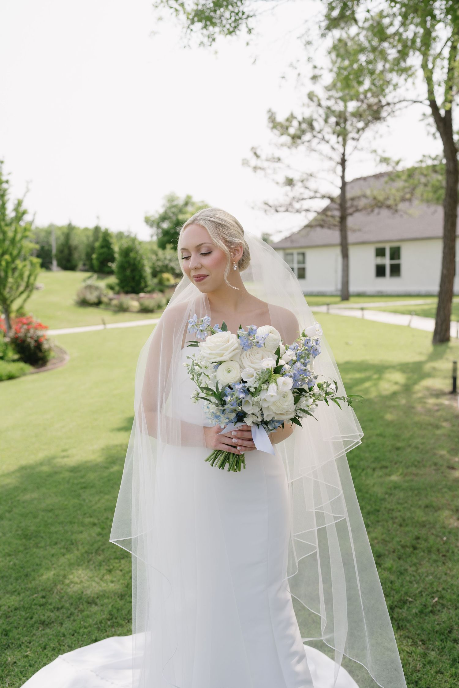 Bride in white gown and veil holds blue and white bouquet outdoors on green lawn near white building.