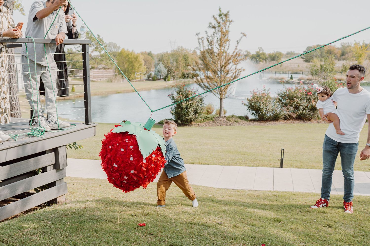 People interact with a swinging red heart decoration on a grassy slope by a lake.