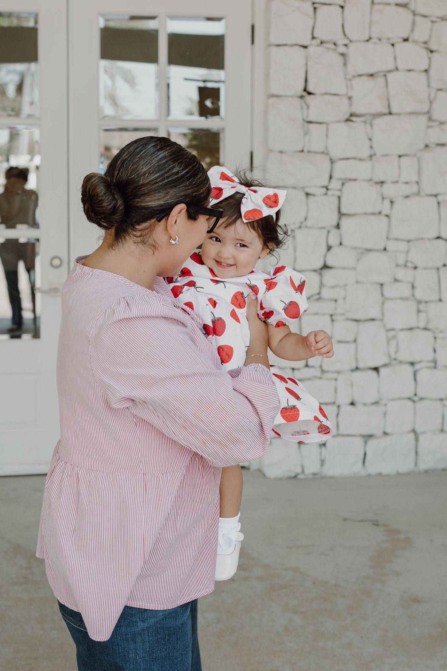 Parent in pink sweater holds child wearing heart-print dress against stone backdrop.