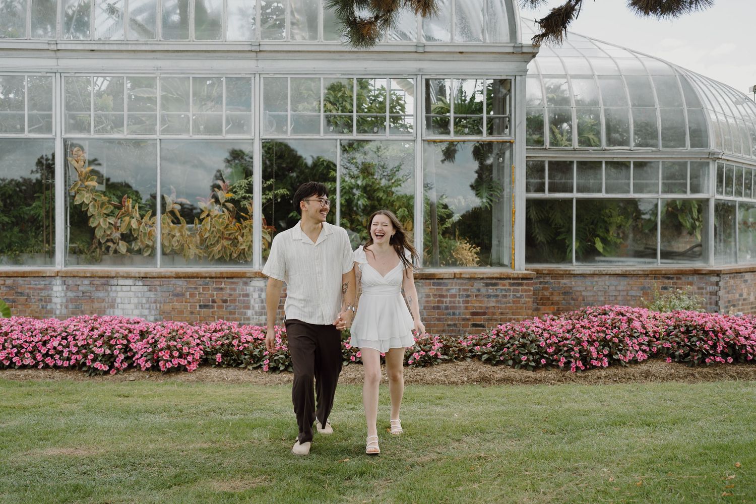 A romantic photoshoot scene outside a large glass conservatory with blooming flower beds.