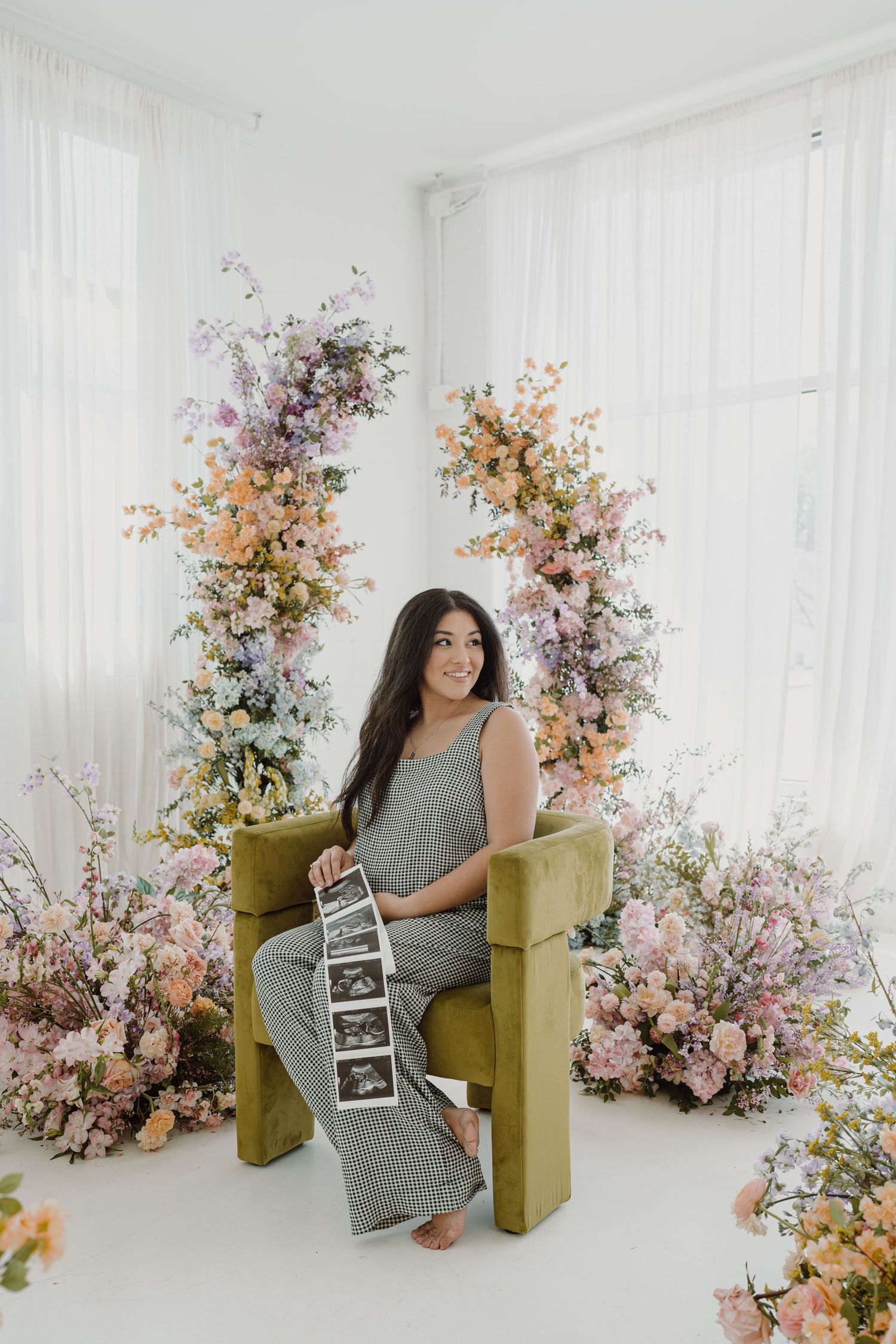 Expectant mother displays ultrasound strip while sitting in yellow chair surrounded by flowers.