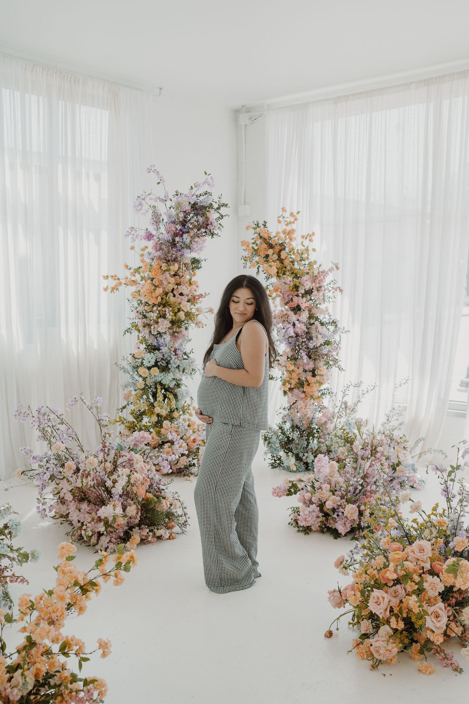 Side-angle maternity photo with romantic floral arrangements and sheer white curtains.
