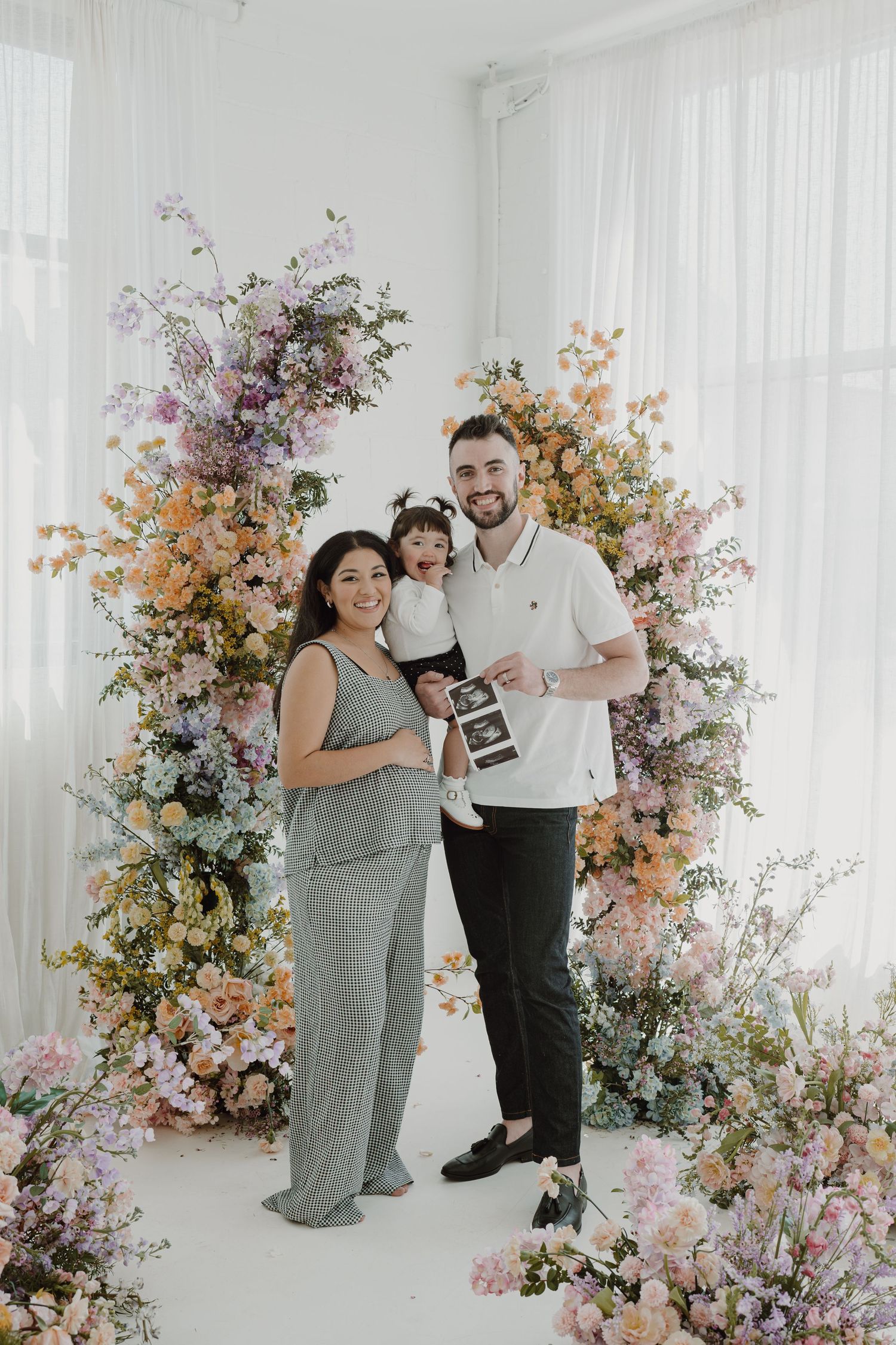 Family pregnancy announcement photoshoot with floral arrangements and ultrasound photos.