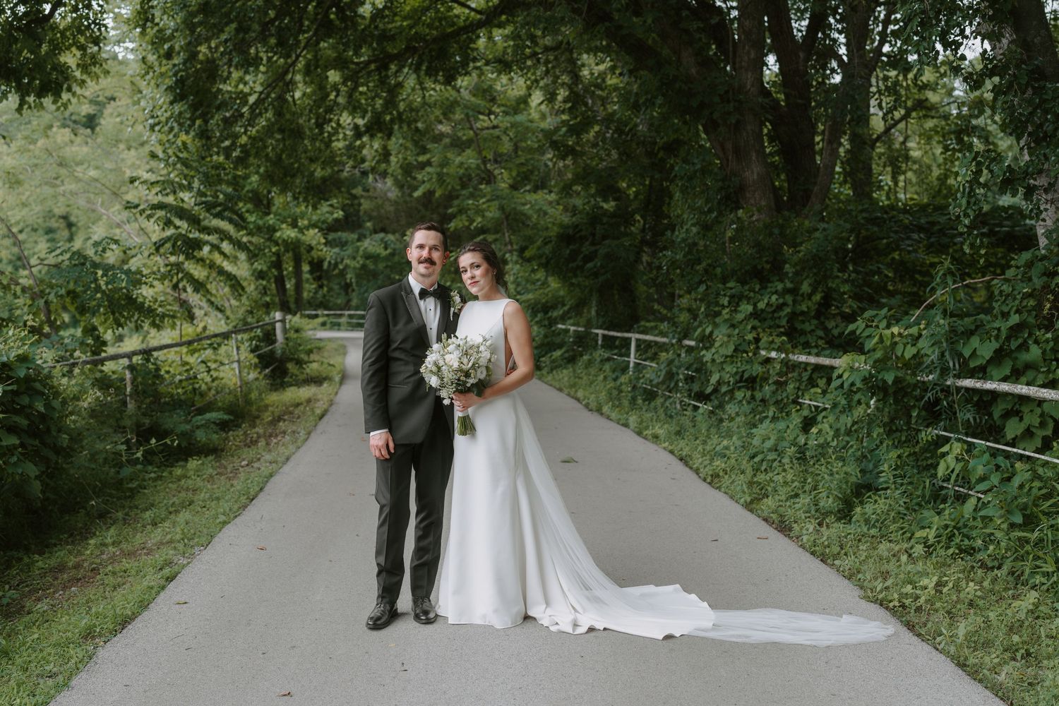 A wedding couple walks together on a tree-lined path in formal attire with lush greenery in the background.