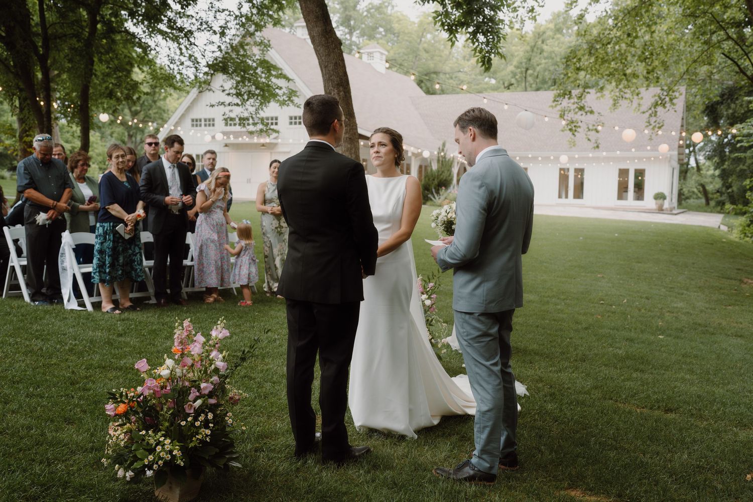 Wedding ceremony in progress on a lush green lawn with guests seated in white chairs.
