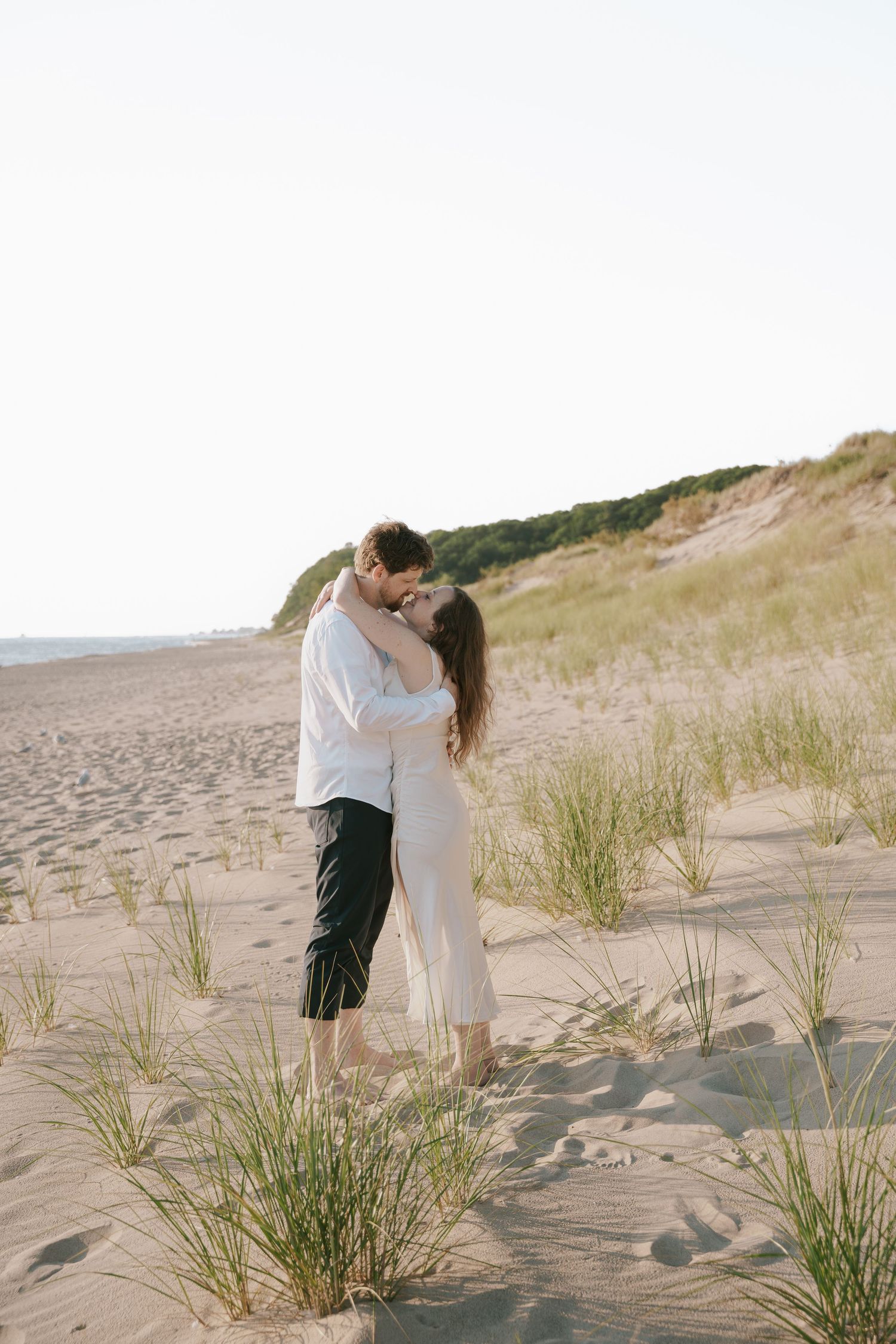 A romantic moment captured between a couple on a sandy beach with dunes and coastal vegetation.