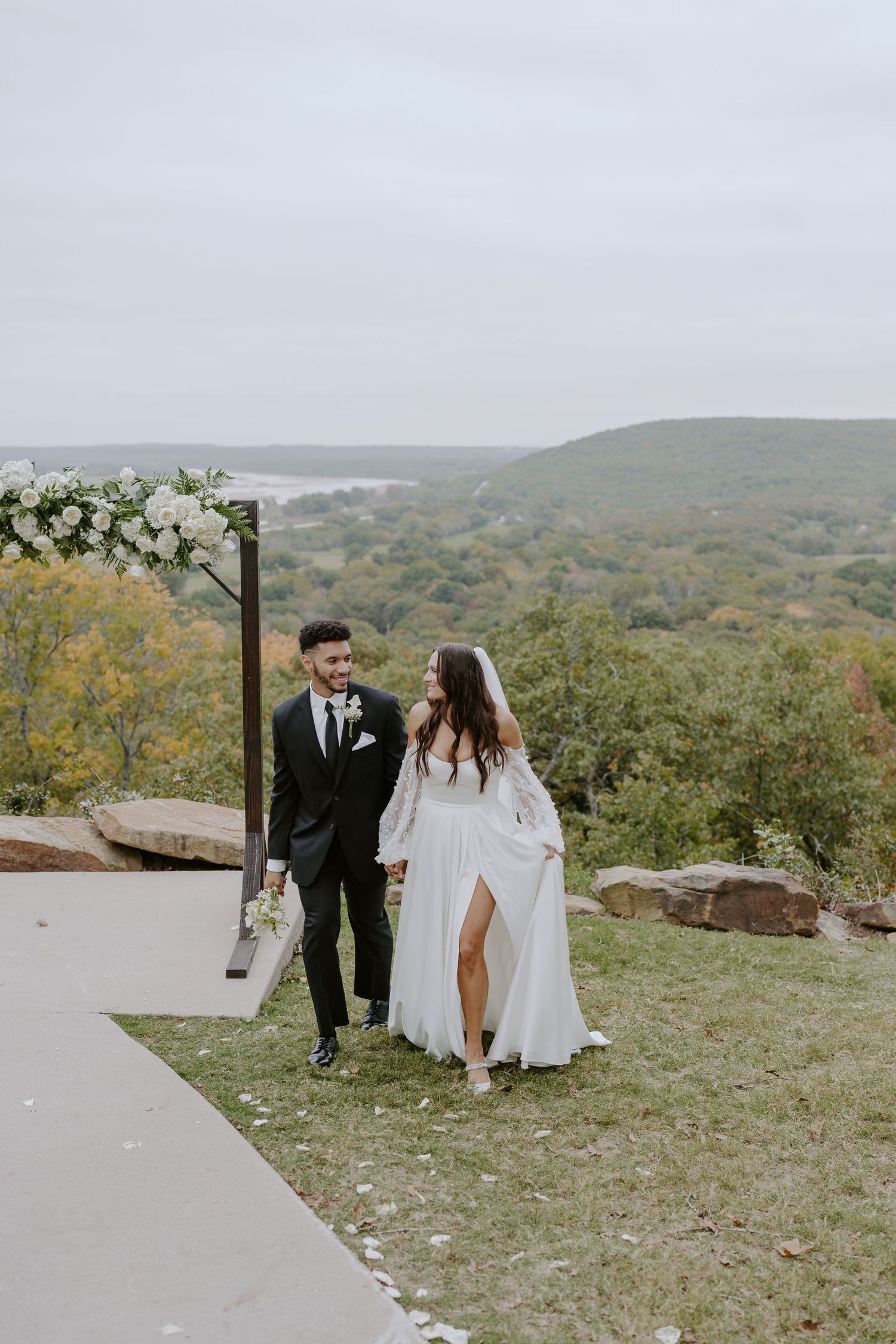 A bride and groom walk together on a scenic mountain path decorated with white floral arrangements.