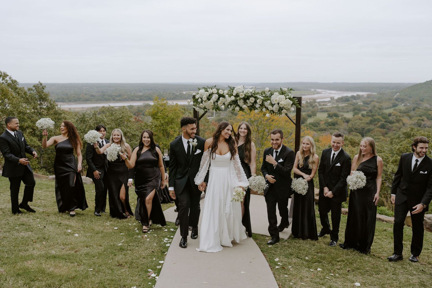 Outdoor wedding scene with white aisle runner and floral arch against rolling hills landscape.