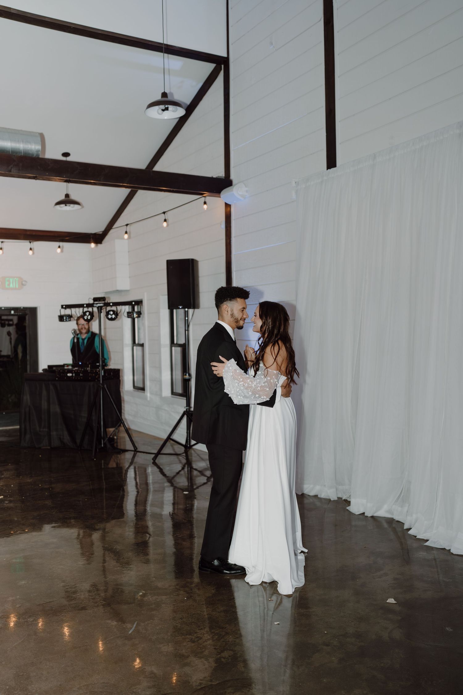 A couple shares a romantic first dance in a modern industrial venue with white drapes and ambient lighting.