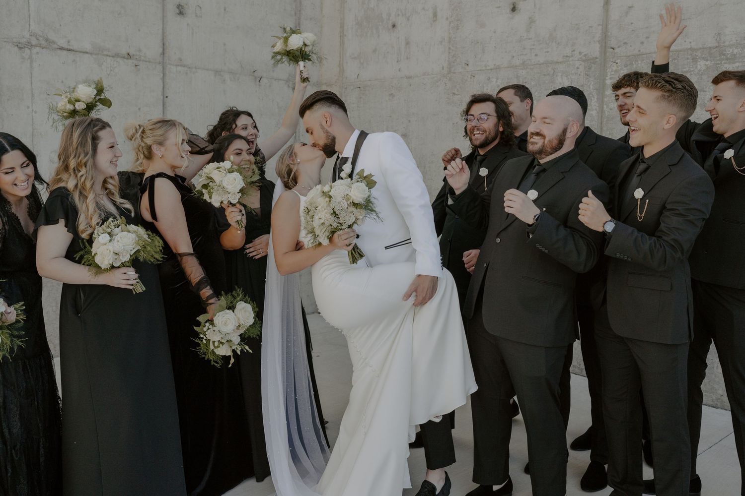 Wedding party in black and white attire celebrates joyfully against concrete wall.