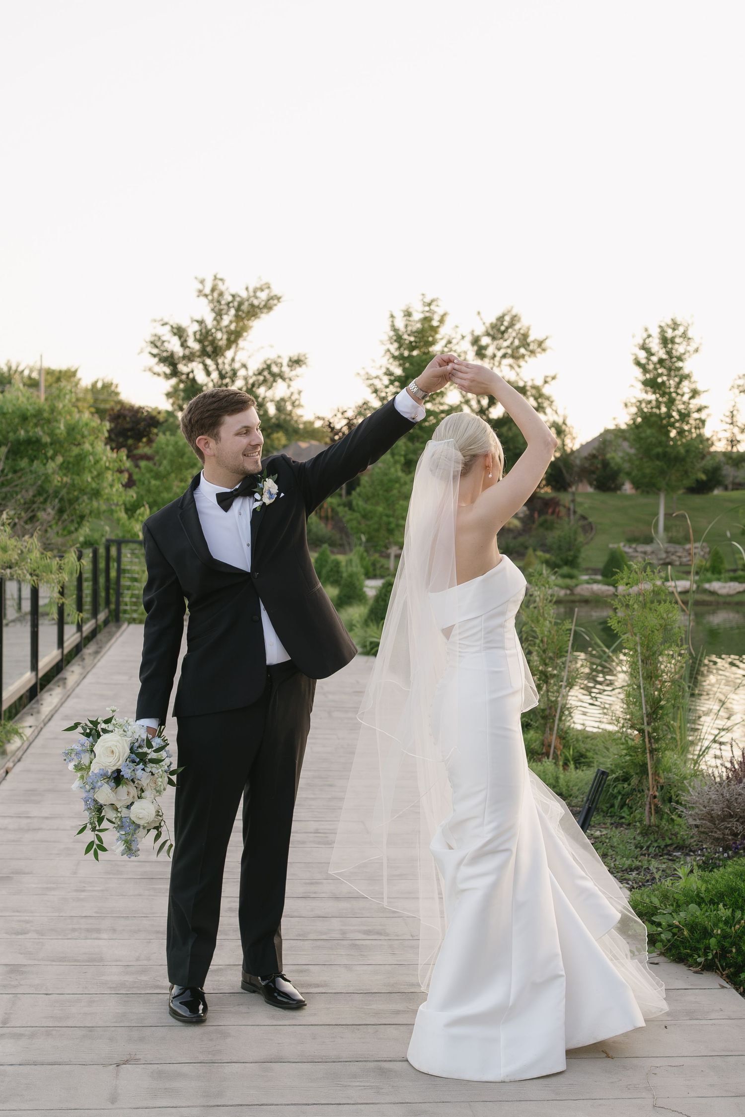 A groom in a black tuxedo twirls his bride in a white gown and veil on an outdoor path at golden hour.