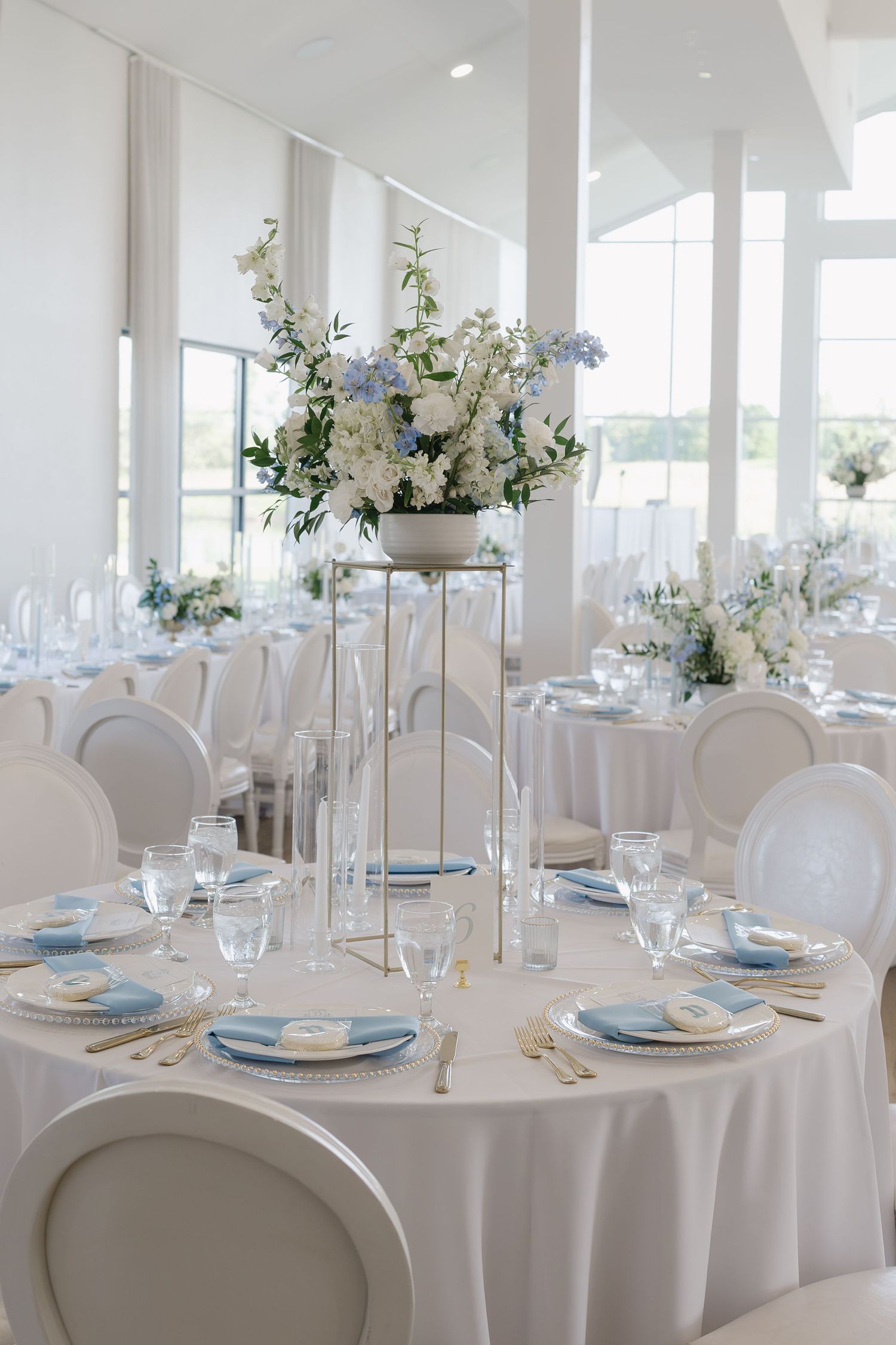 Elegant white wedding reception hall with blue floral centerpieces, white chairs, and blue-accented table settings.