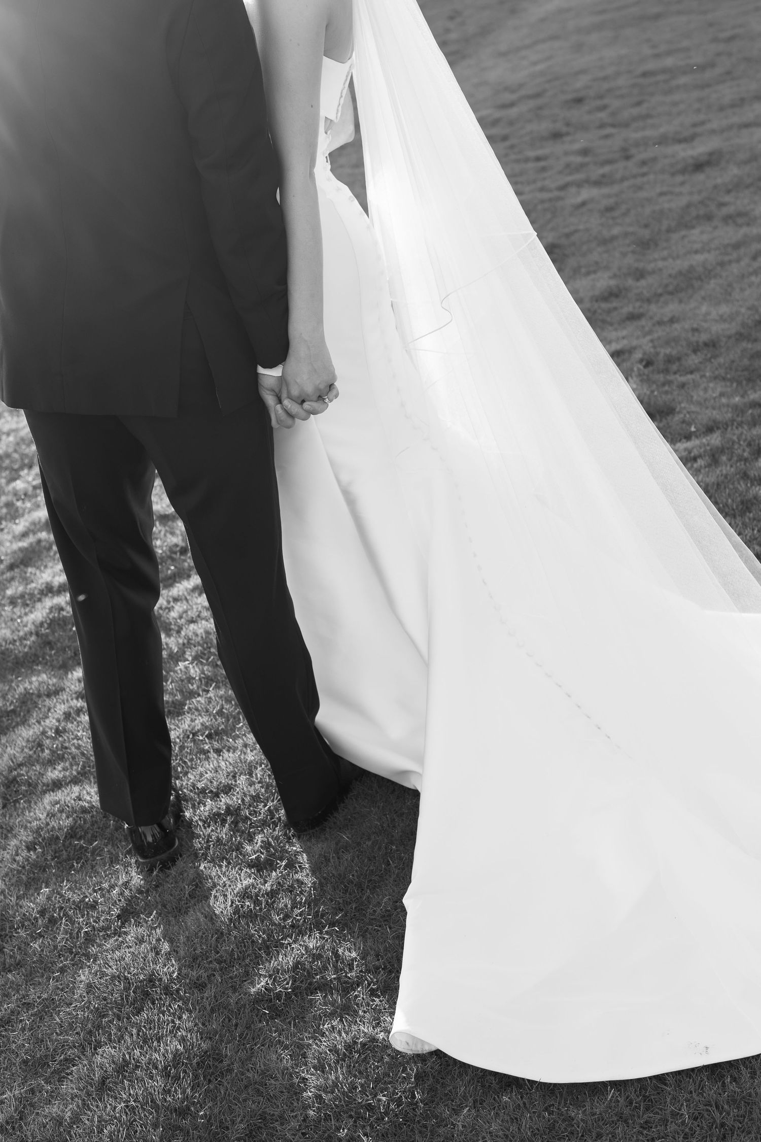 Black and white photo of bride and groom holding hands, walking on grass, with flowing wedding dress train.
