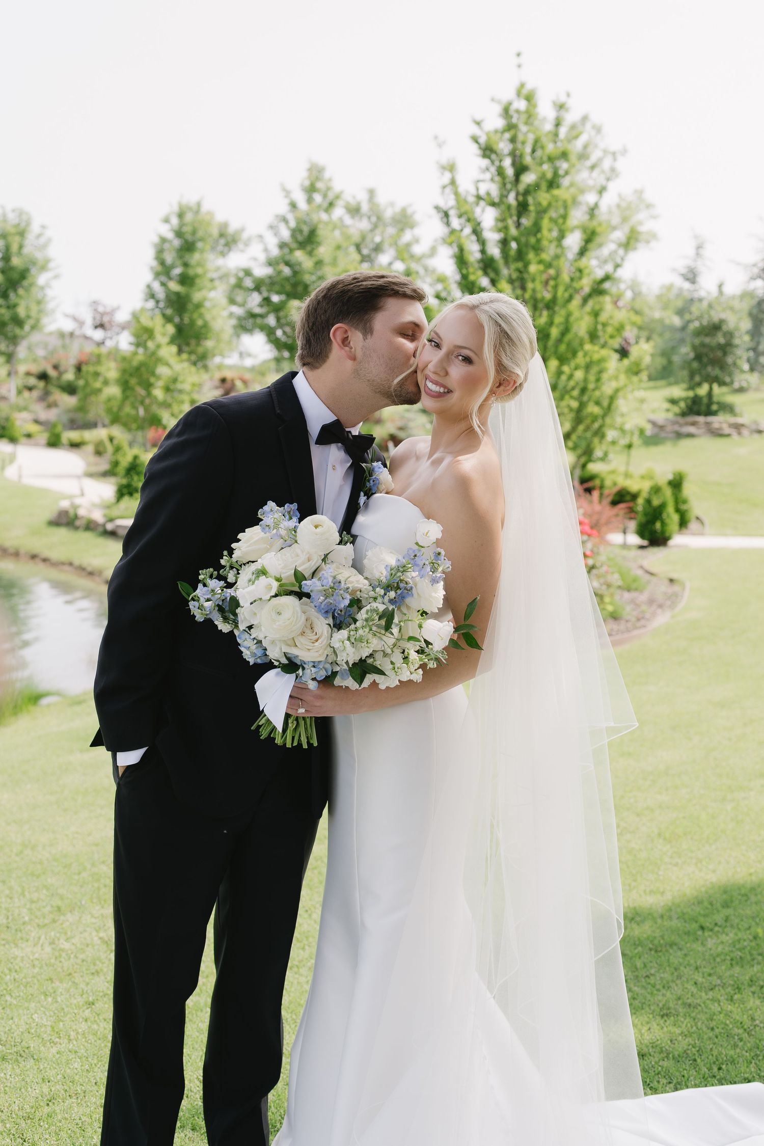Groom kisses bride on cheek as she smiles holding white and blue bouquet, standing on green lawn at outdoor wedding.