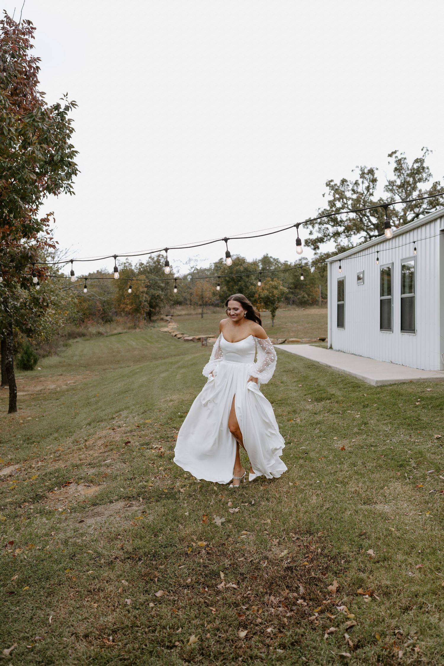 Person in off-shoulder white wedding gown twirls on lawn with string lights and white building in background.