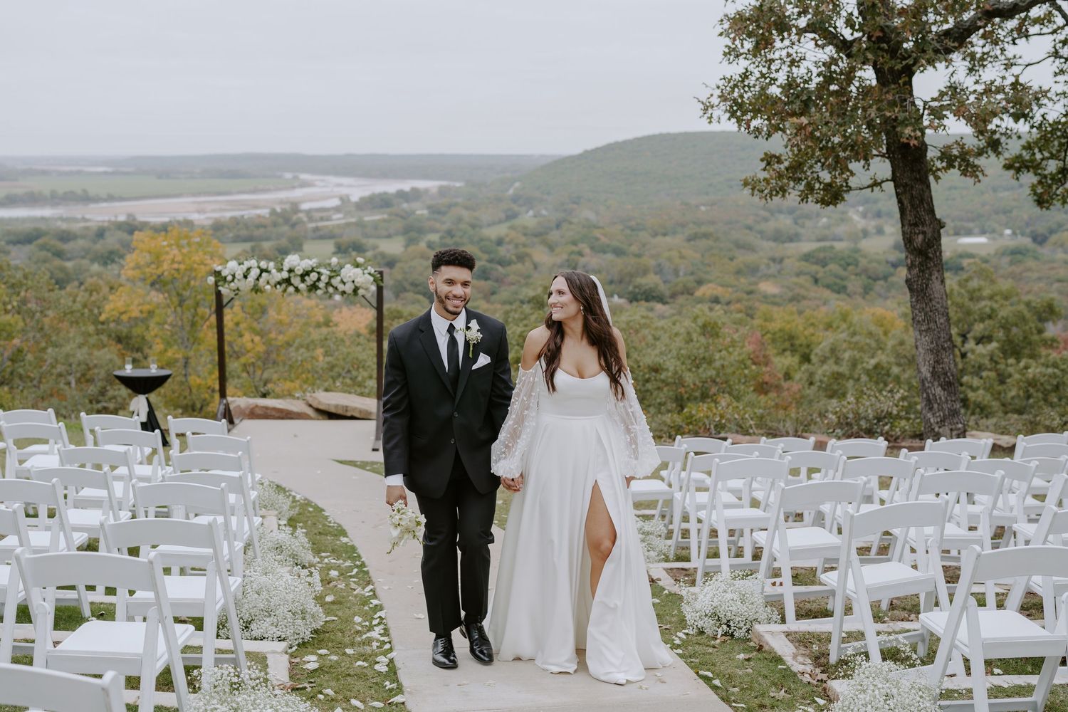 An outdoor wedding venue setup with white chairs and floral decorations on a scenic mountain overlook.