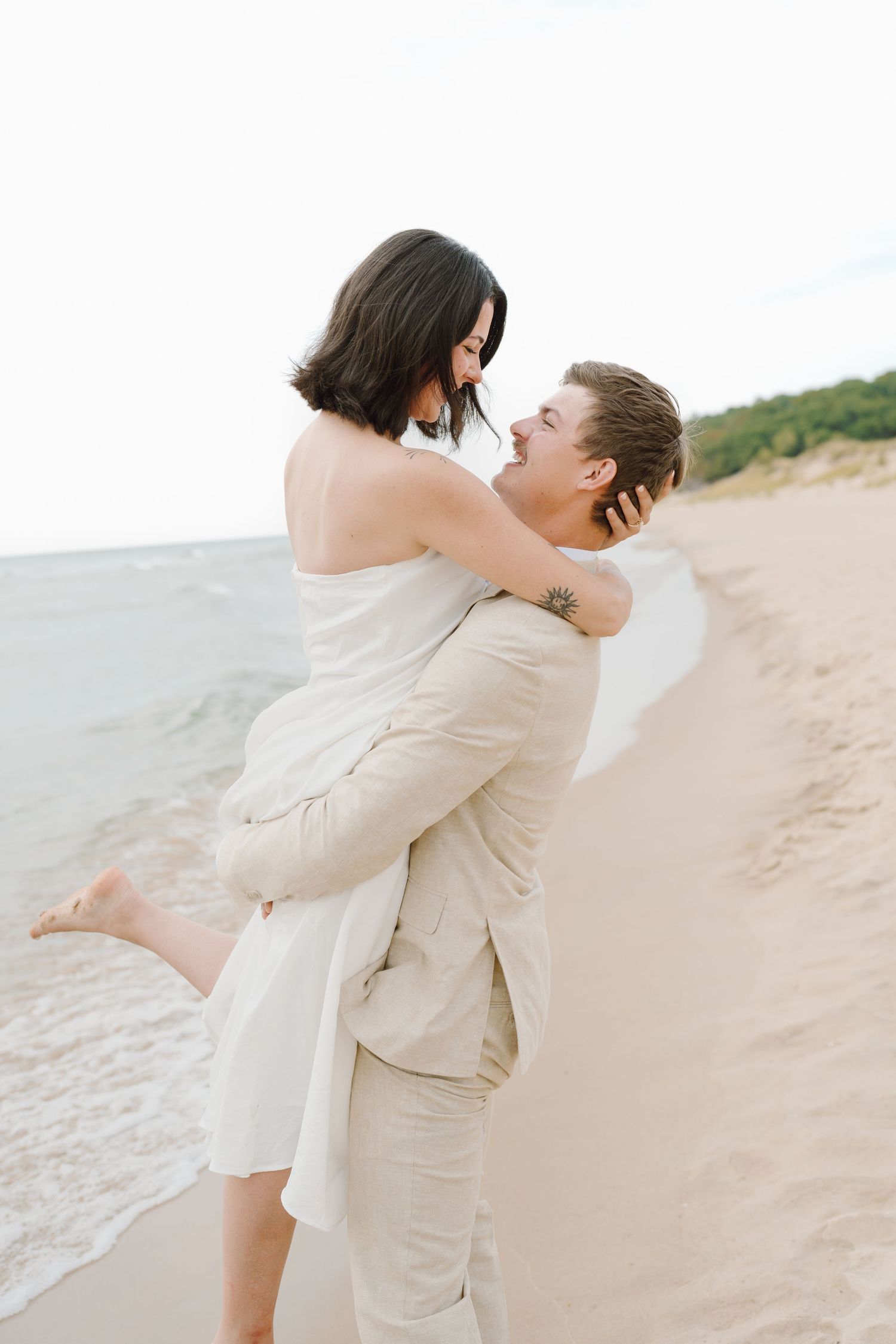 A romantic couple in light colored outfits embrace and kiss on a sandy beach with ocean waves in the background.