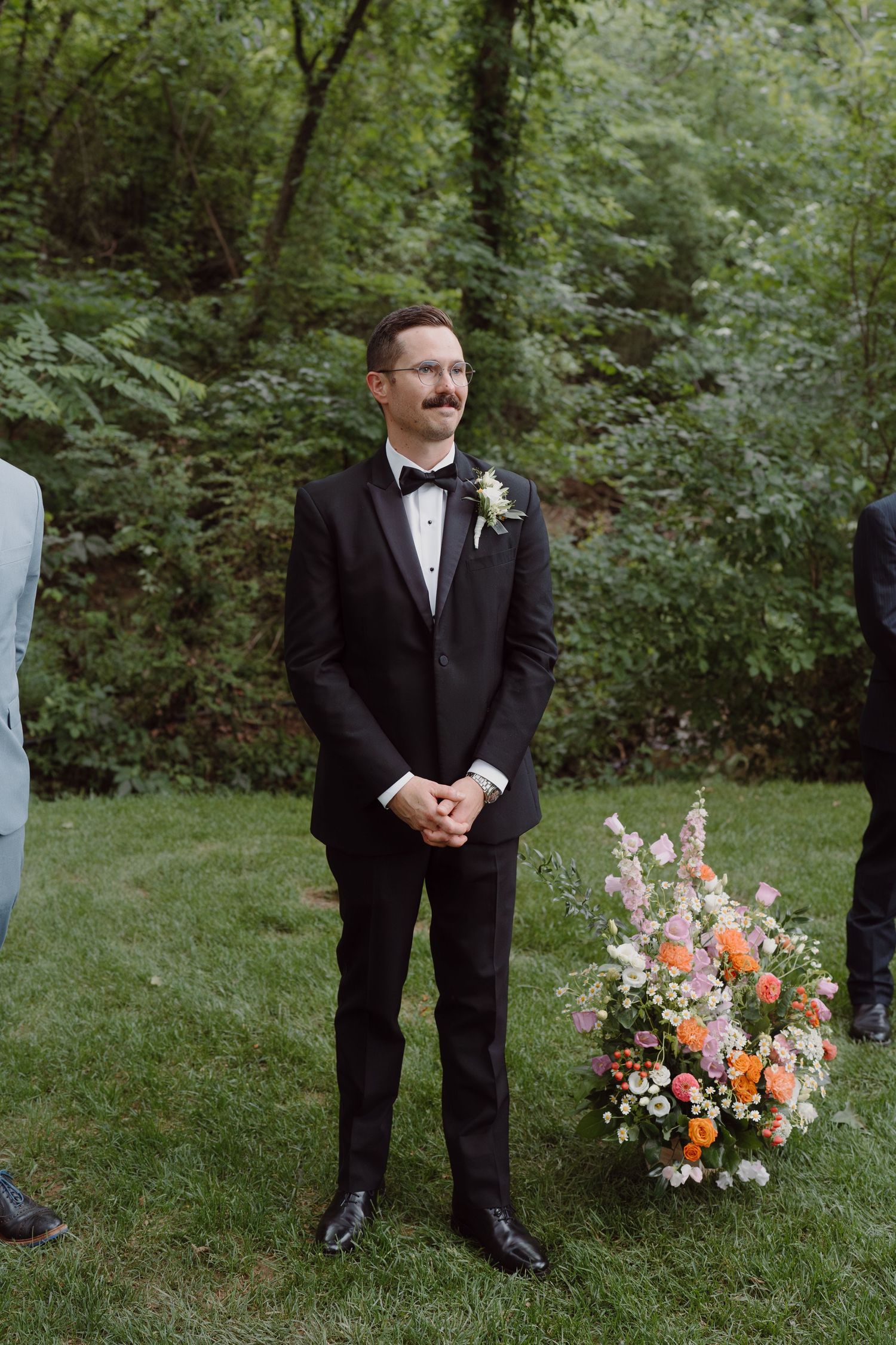 A groom in a black tuxedo stands in a wooded outdoor setting with colorful floral arrangements nearby.