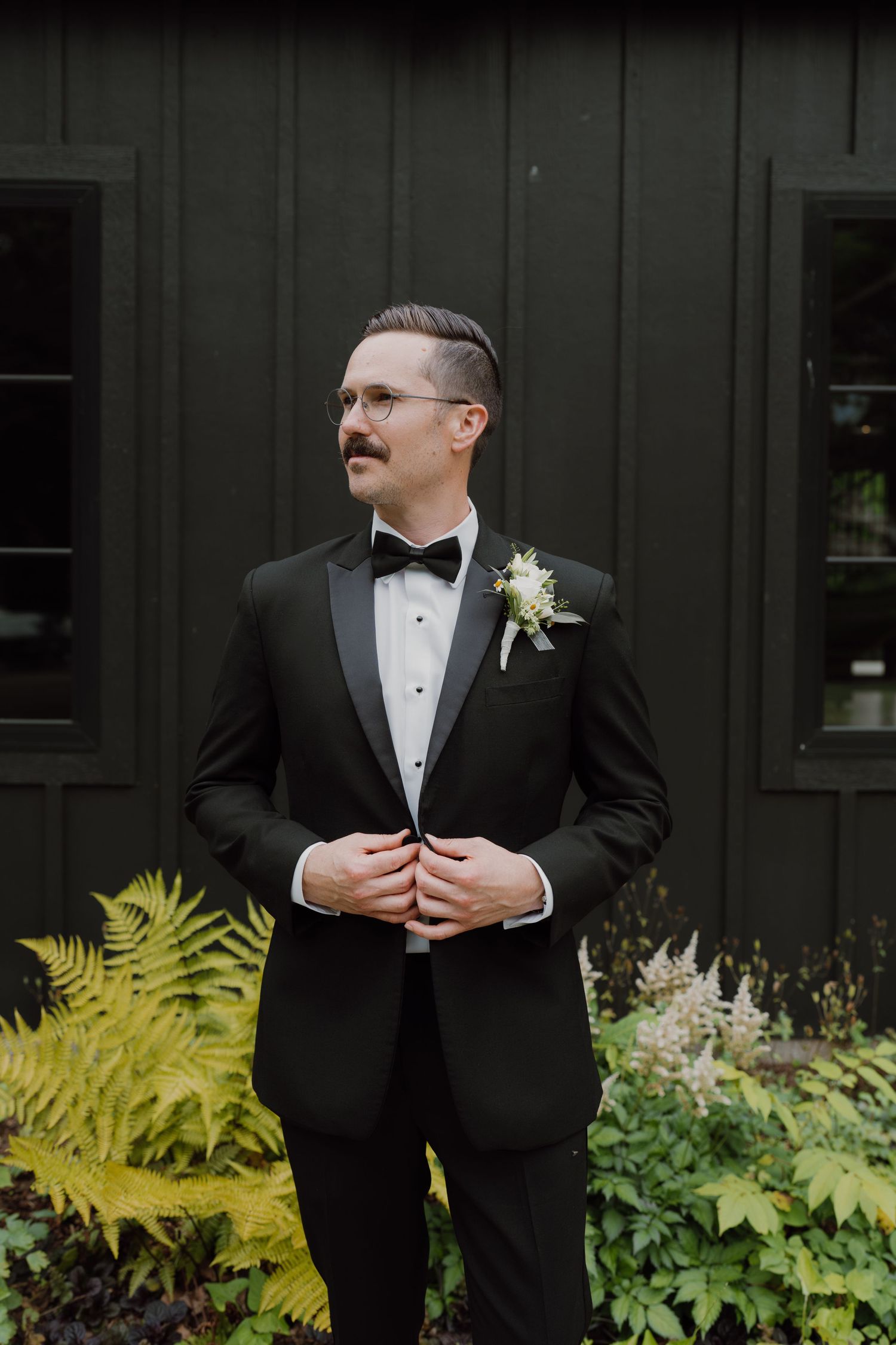 Groom adjusts jacket while standing against dark wall with vibrant garden foliage.