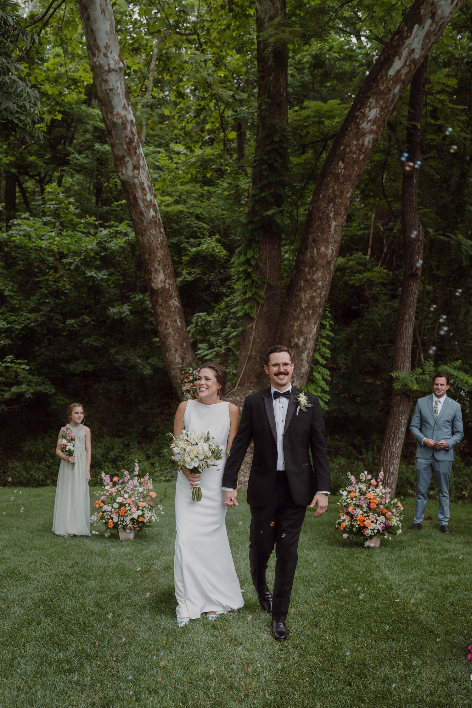 Wedding party members walking through a forest clearing with floral decorations and scattered guests.