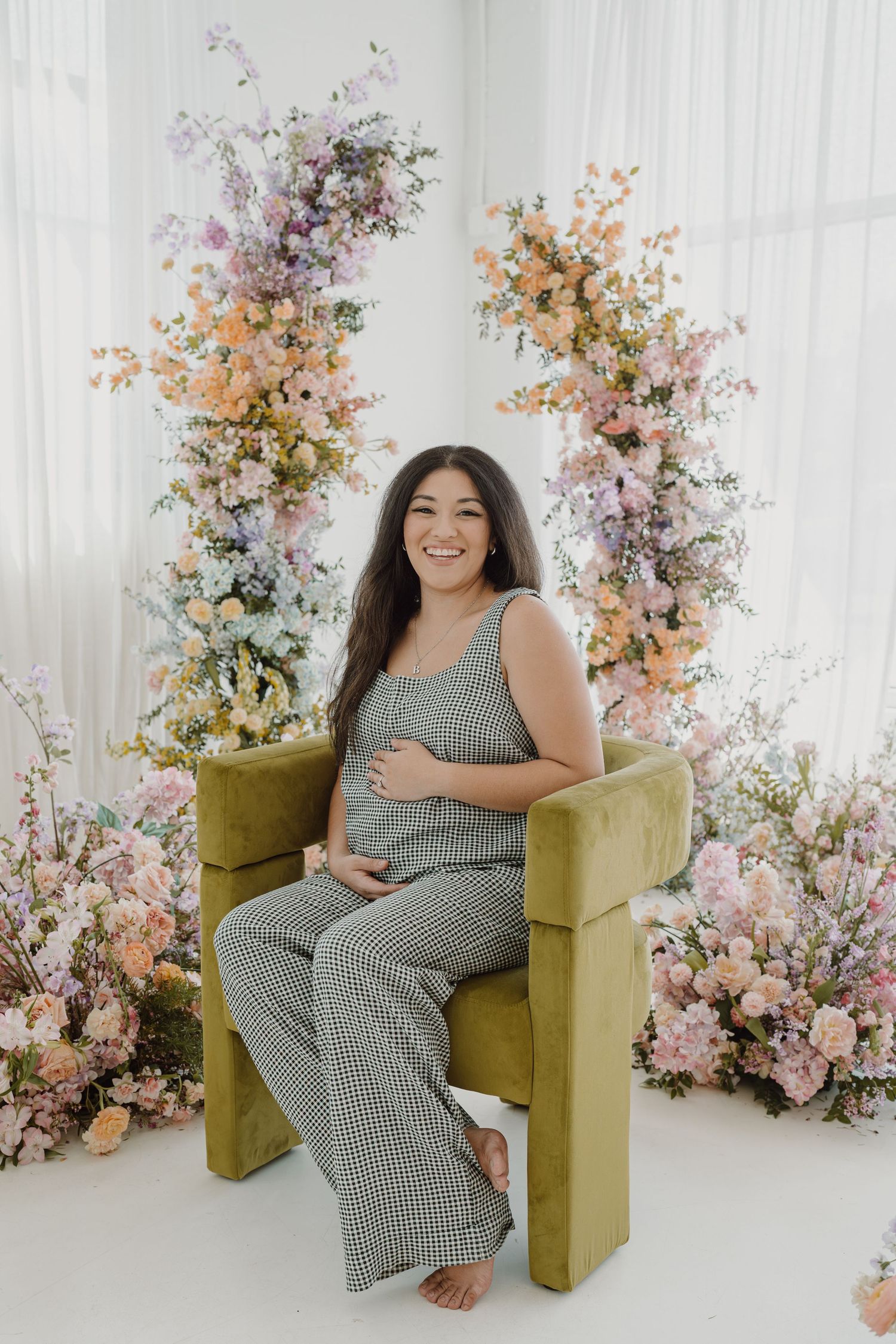 Expectant mother poses on mustard velvet chair surrounded by pink and peach floral arrangements.