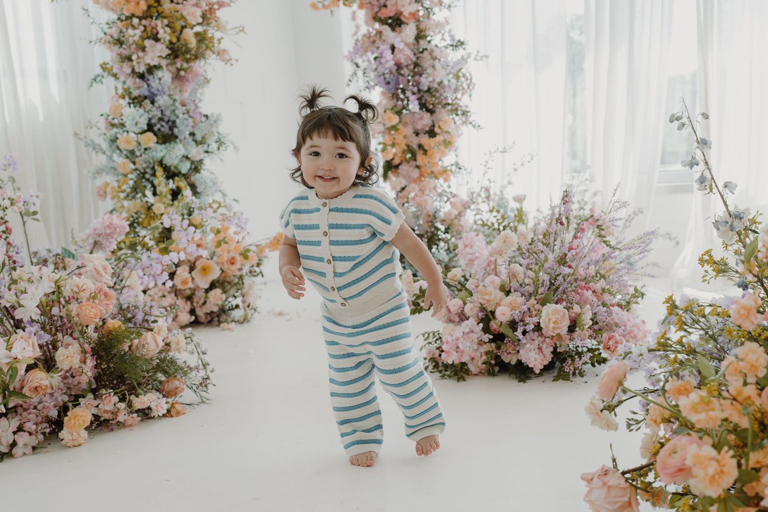 Energetic toddler jumping in striped outfit surrounded by beautiful floral arrangements.