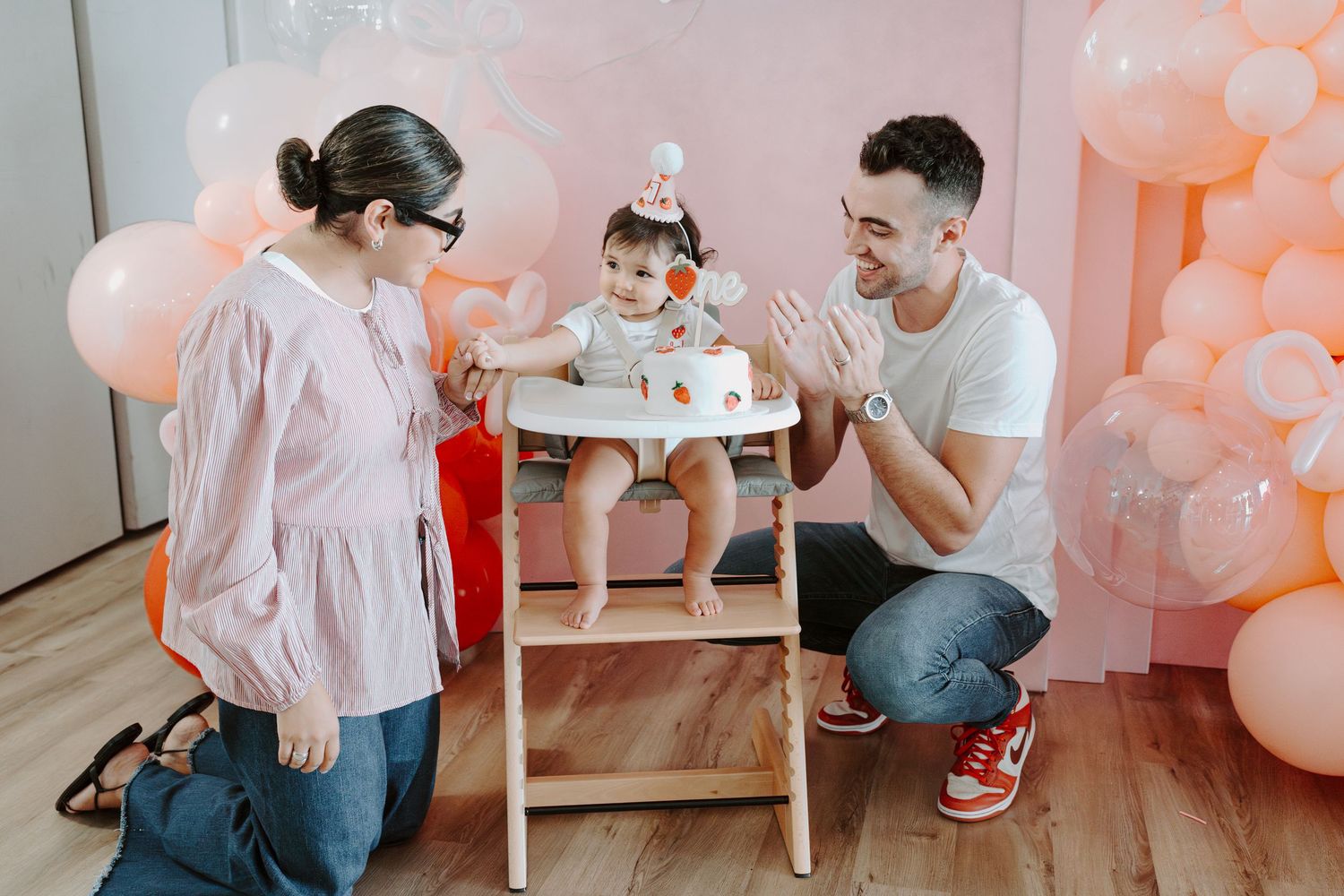 A birthday celebration with a baby in a high chair surrounded by pink balloons and decorations.